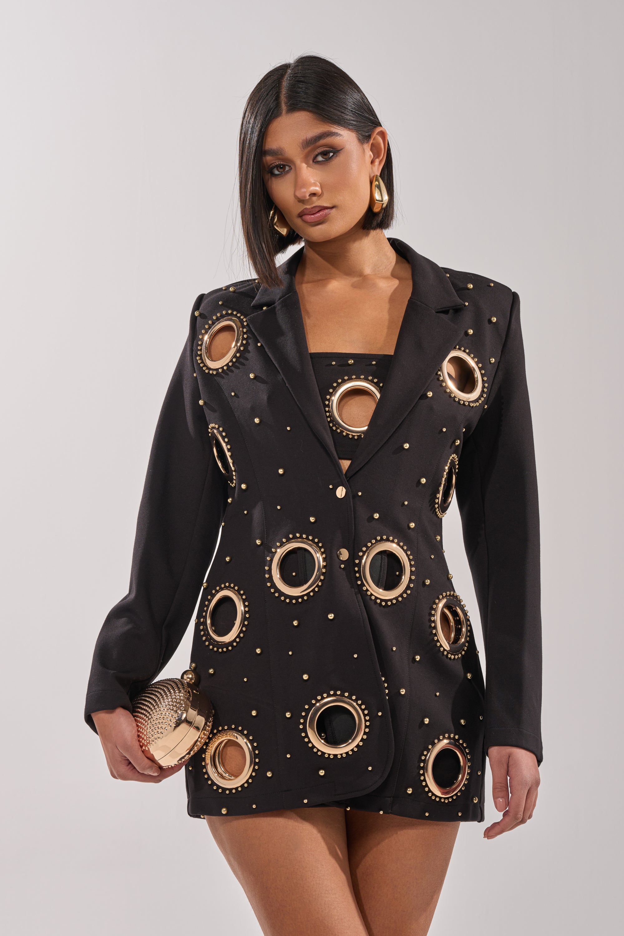 A woman wears the SHOW STOPPER BLAZER with gold studs and circular cutouts over a matching top, accessorized with a gold textured clutch and earrings. Her straight dark hair is styled to one side. 3