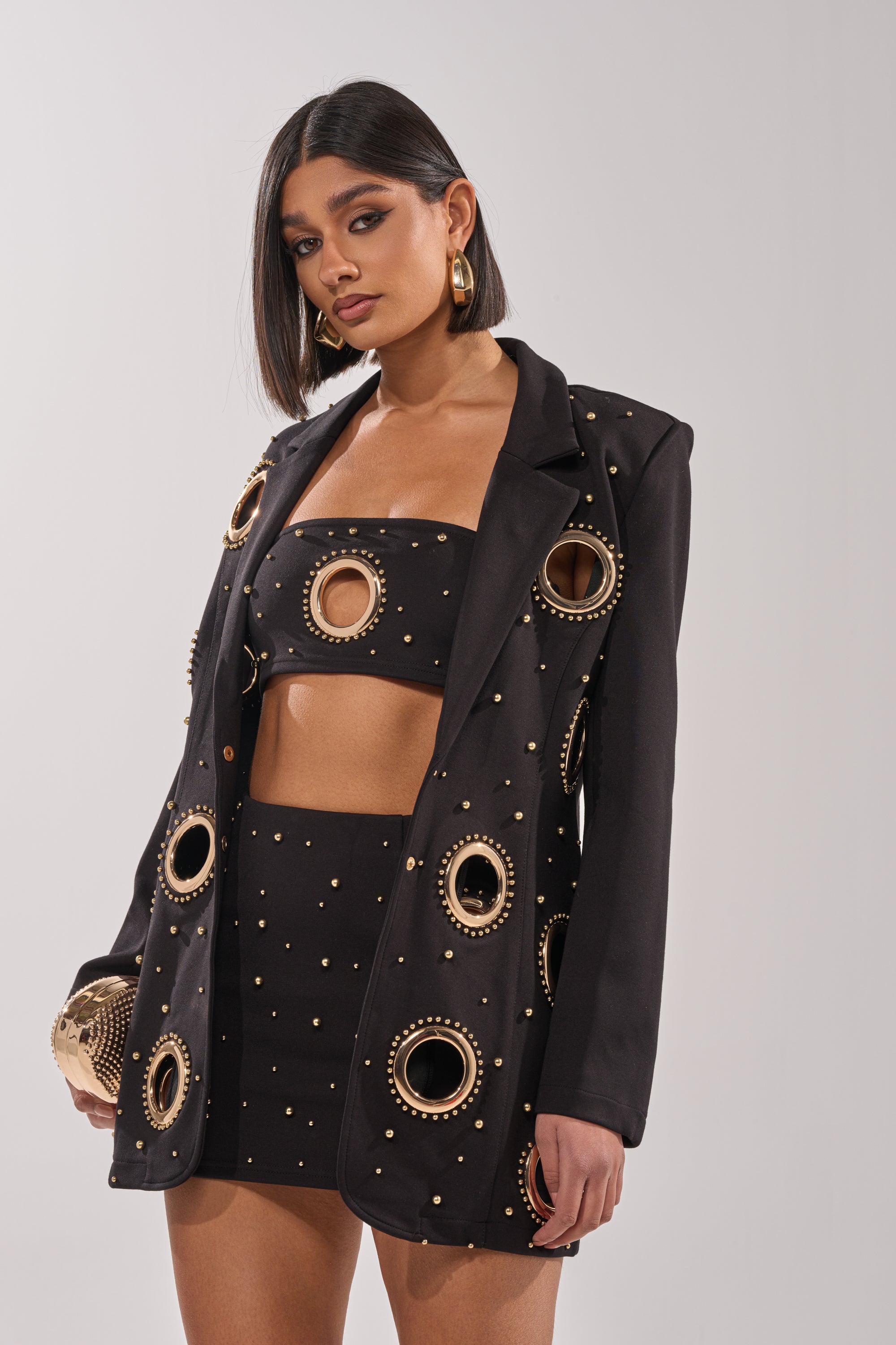 A woman with a sleek bob wears the SHOW STOPPER BLAZER, featuring gold studs and bold cutouts, styled with hoop earrings and a gold clutch. 4