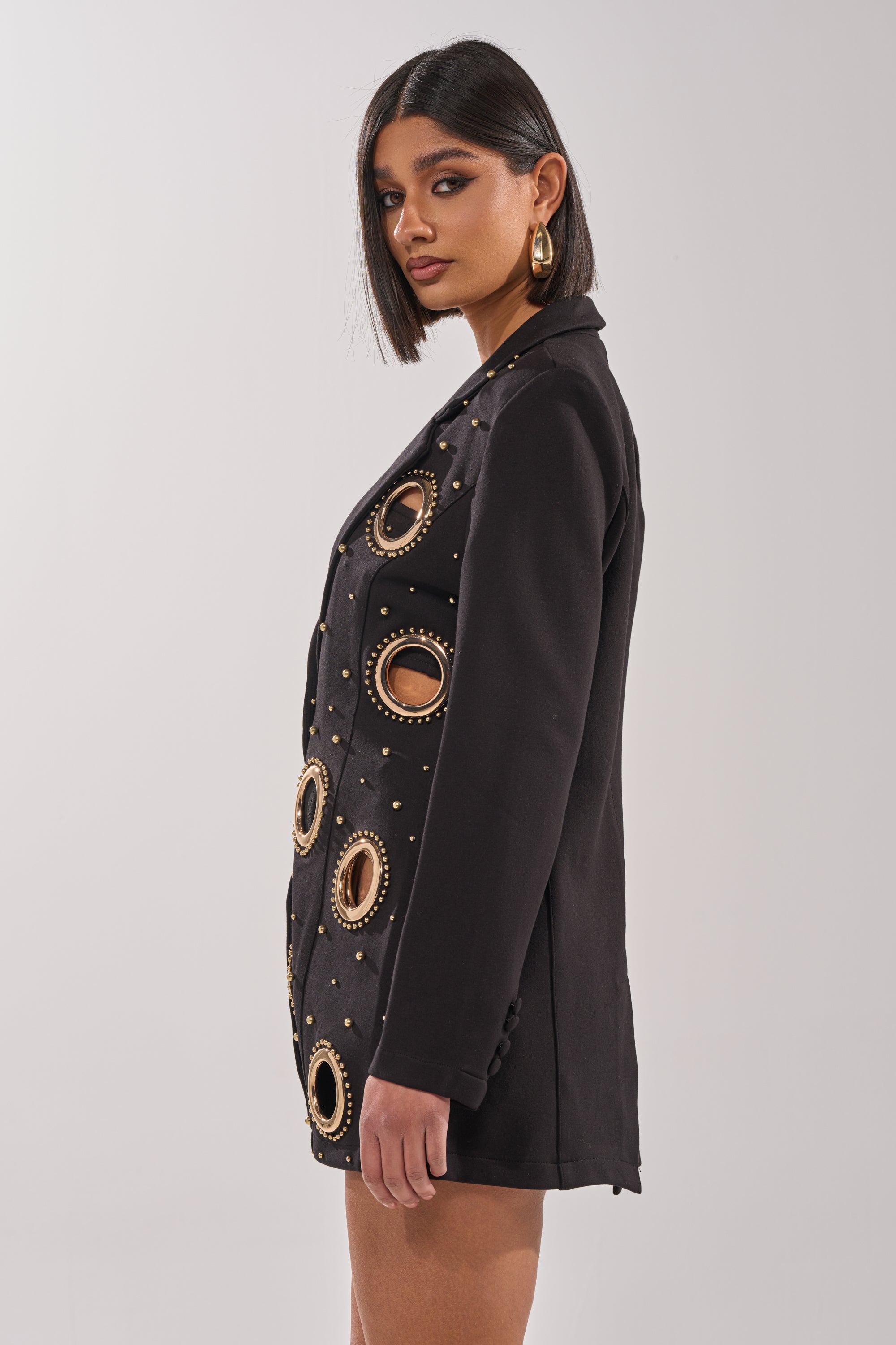 A woman with a sleek bob models the SHOW STOPPER BLAZER, styled with a tailored fit and gold-tone dome studs, posing in profile against a plain light background. 5