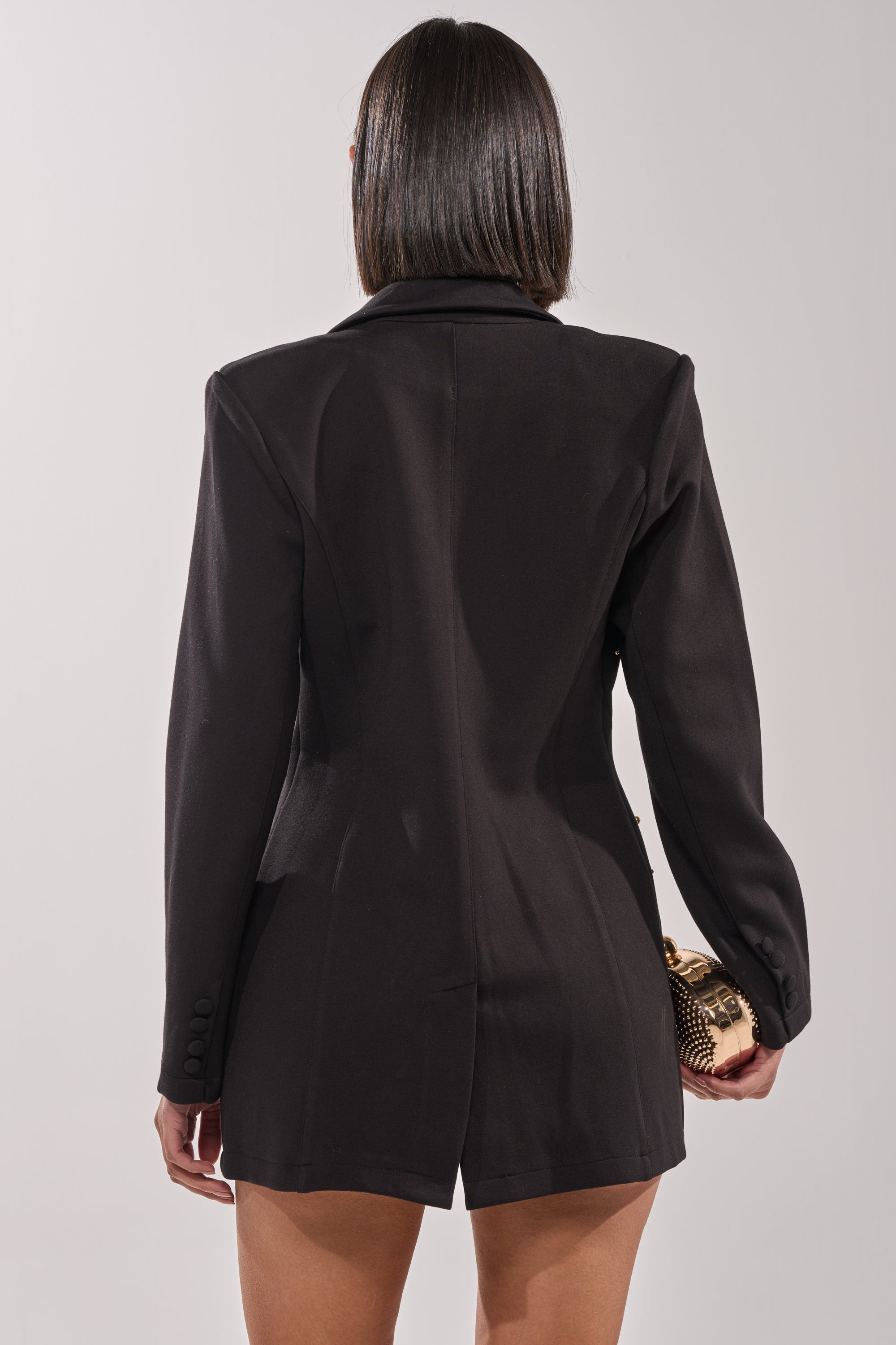 A person with straight, shoulder-length dark hair is shown from behind, wearing the SHOW STOPPER BLAZER and holding a small gold clutch against a plain, light-colored background. 6