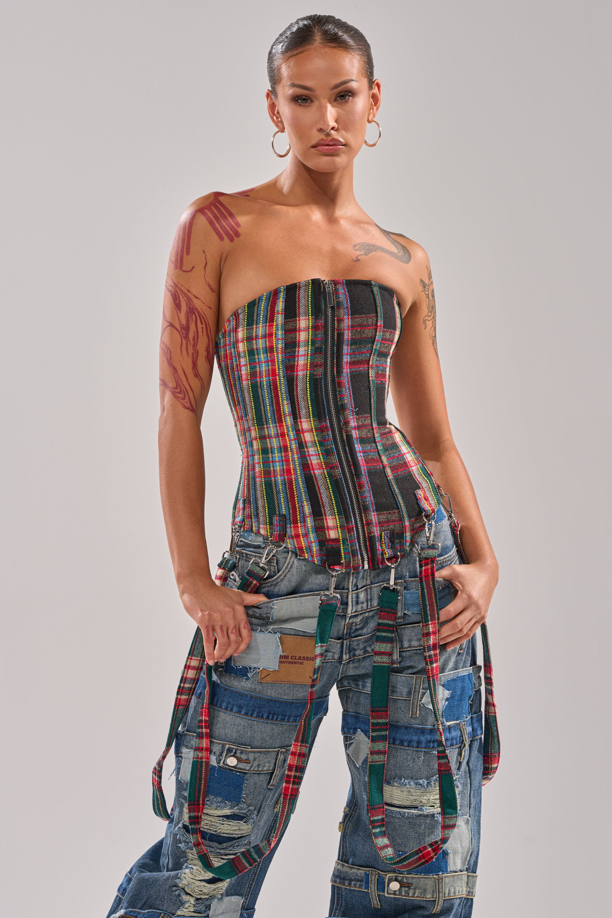 Wearing the TALK TO ME NICE PLAID CORSET and patchwork denim jeans with suspenders, a person stands with hands on hips, accessorized with hoop earrings, tattoos, and slicked-back hair.
