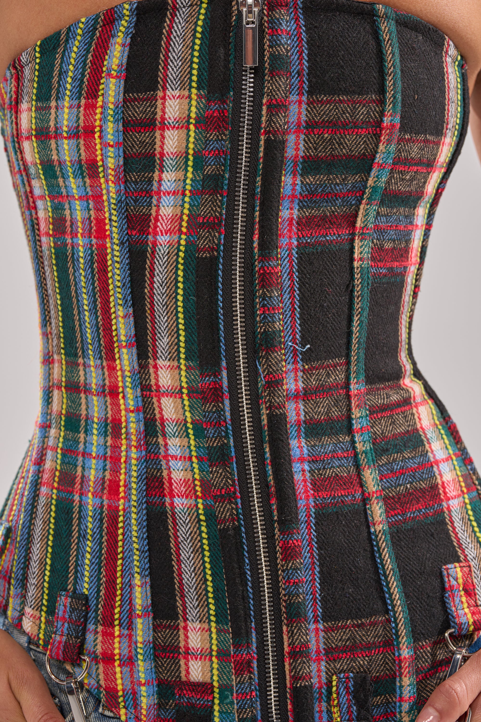 Close-up of a person wearing the TALK TO ME NICE PLAID CORSET featuring a central zipper, bottom buckle details, and vibrant red, green, yellow, blue, and black tartan patterns against a light background.