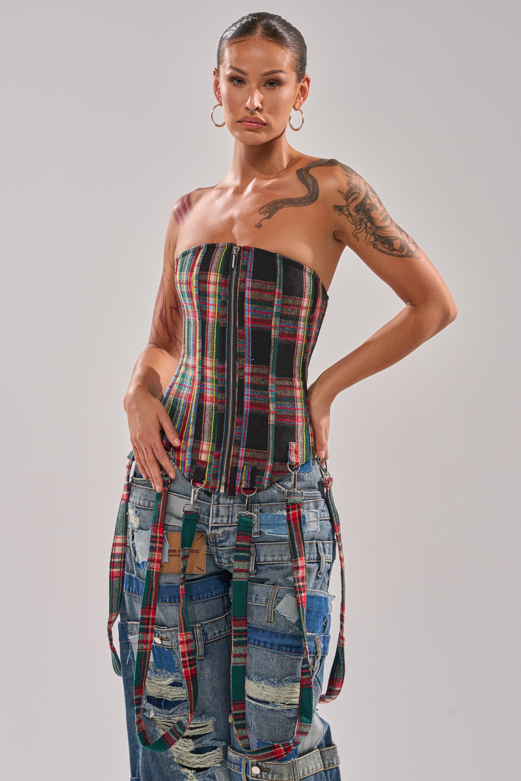 A woman with hoop earrings wears the TALK TO ME NICE PLAID CORSET with detachable garter straps and patchwork denim jeans featuring plaid accents. She poses against a plain light background, showing off arm tattoos and slicked-back hair.