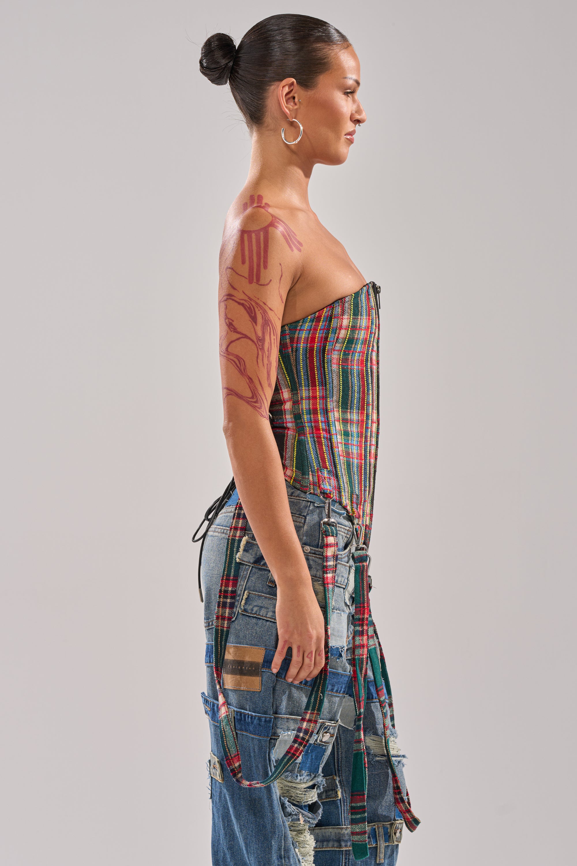 A woman in profile with a slicked-back bun and hoop earrings wears the TALK TO ME NICE PLAID CORSET, patchwork denim pants, and shows a bold red abstract tattoo on her upper arm. The background is light gray.