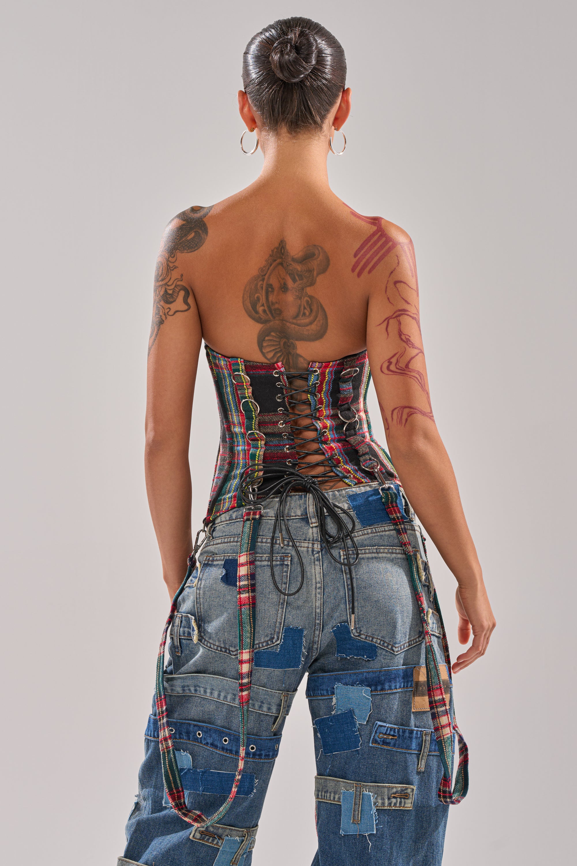 A person stands with their back to the camera, wearing the TALK TO ME NICE PLAID CORSET with a lace-up back and patchwork denim jeans. Their hair is in a bun, and tattoos are visible on their back and arm.