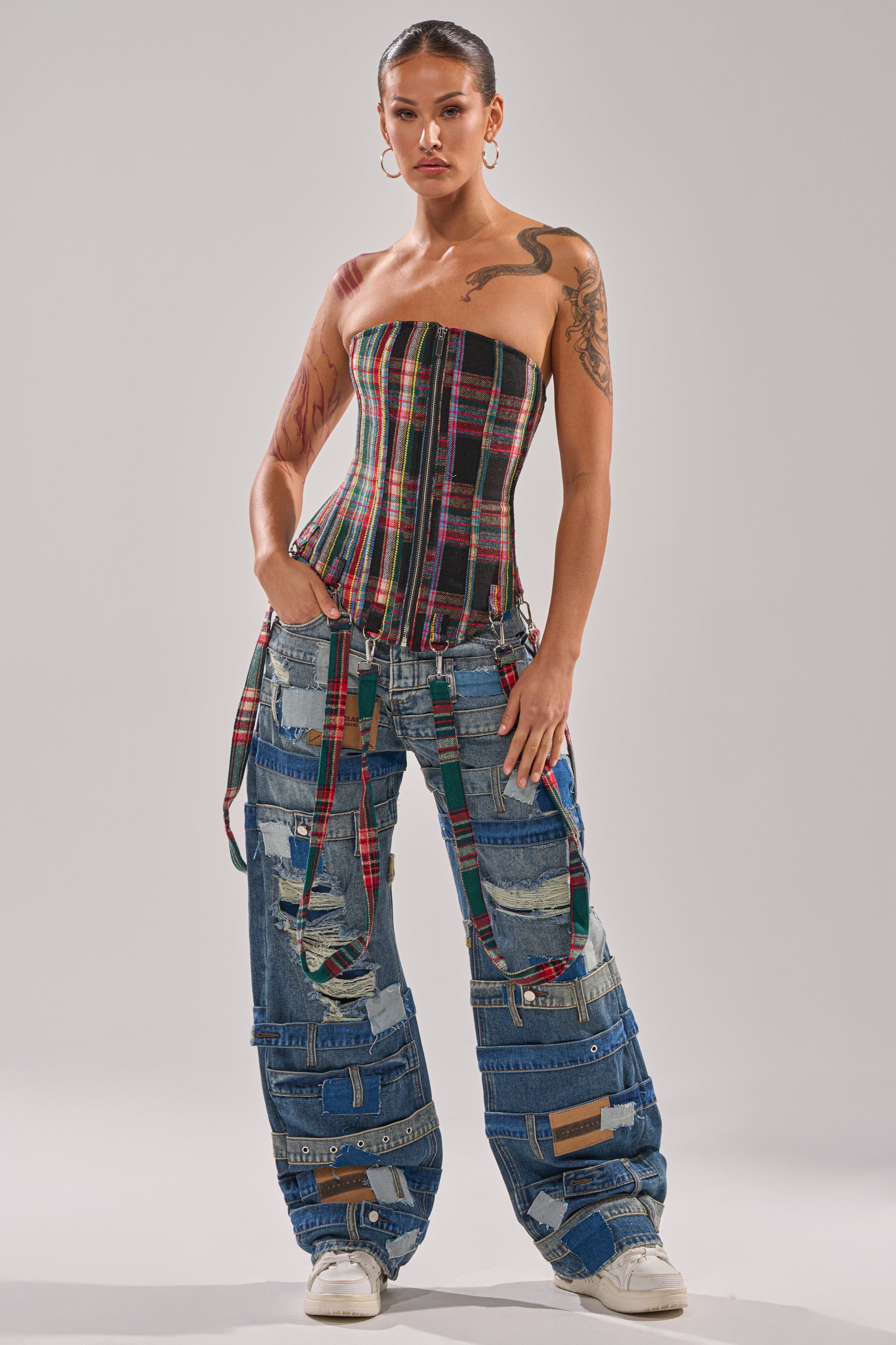 A woman stands confidently in the TALK TO ME NICE PLAID CORSET, paired with baggy, patchwork denim jeans in various blue shades and distressed textures. She completes her look with white sneakers and displays her arm tattoos.