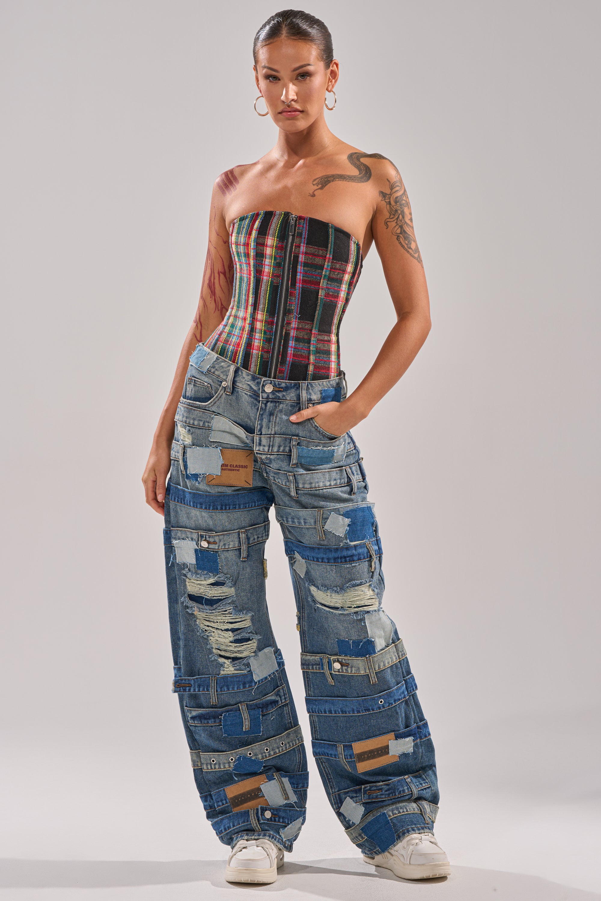 A woman wears the TALK TO ME NICE PLAID CORSET with a lace-up back, paired with loose, patched distressed denim jeans, hoop earrings, white sneakers, and showcases visible tattoos while standing confidently against a plain background.