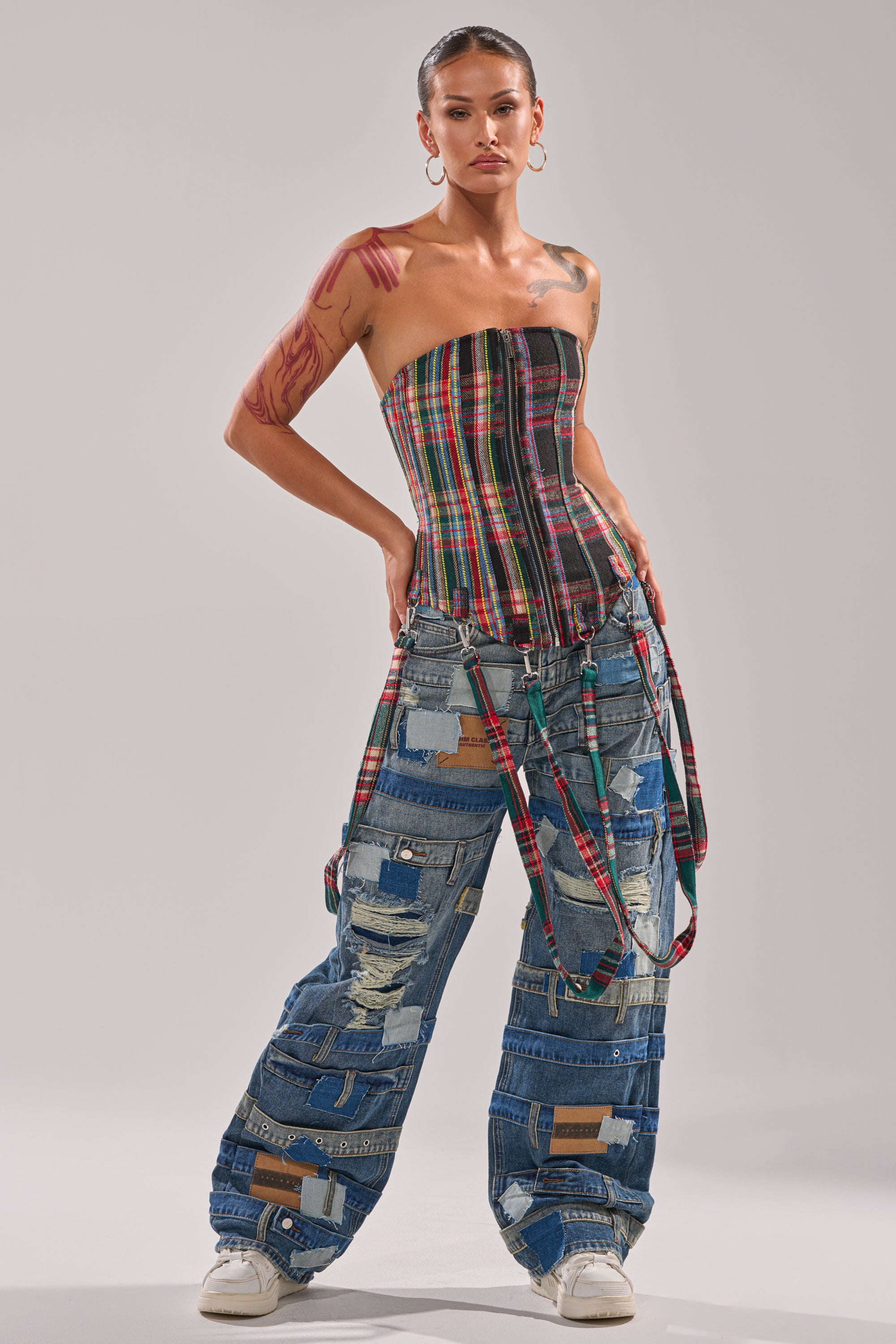 Wearing the TALK TO ME NICE PLAID CORSET, wide-leg patchwork denim jeans with distressed details, and white sneakers, they accessorize with hoop earrings, slicked-back hair, and a bold red arm tattoo.