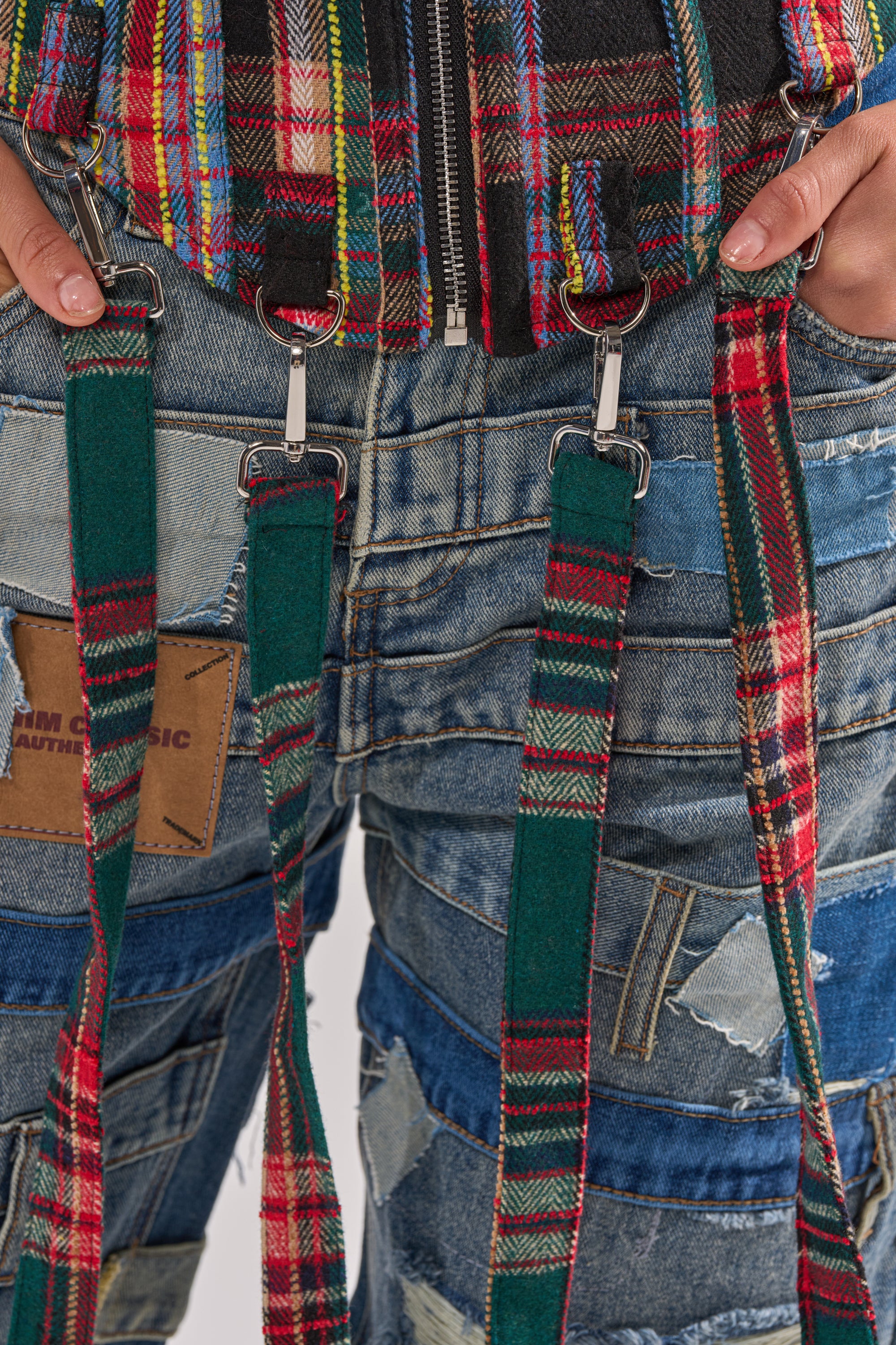 A close-up of a person wearing patched blue jeans and a plaid jacket with the TALK TO ME NICE PLAID CORSET suspenders hanging down, held in their hands.