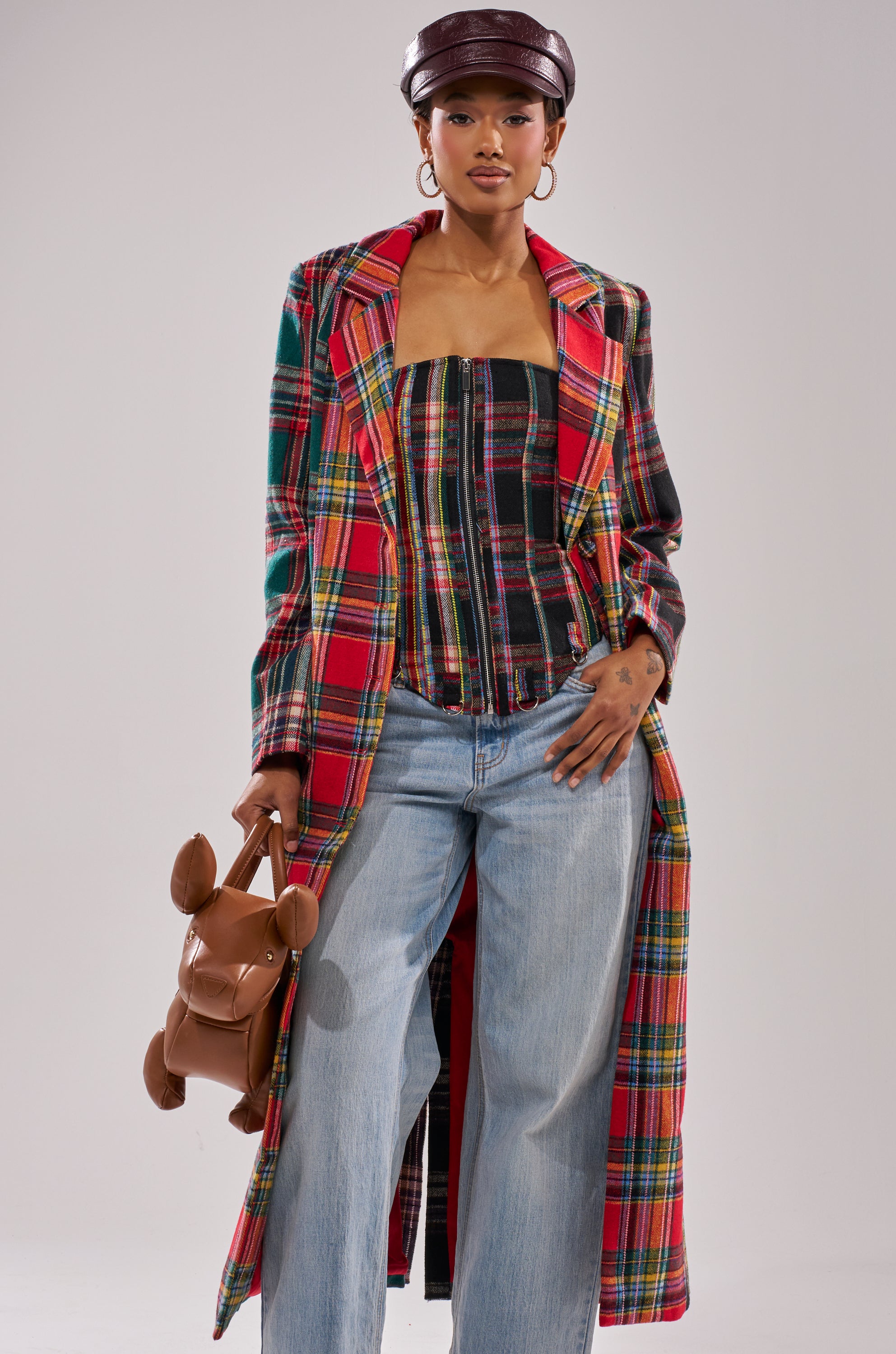 A woman wears the TALK TO ME NICE PATCHWORK TRENCH over a matching top and light blue jeans, accessorized with a brown dog-shaped bag, dark leather cap, and hoop earrings.