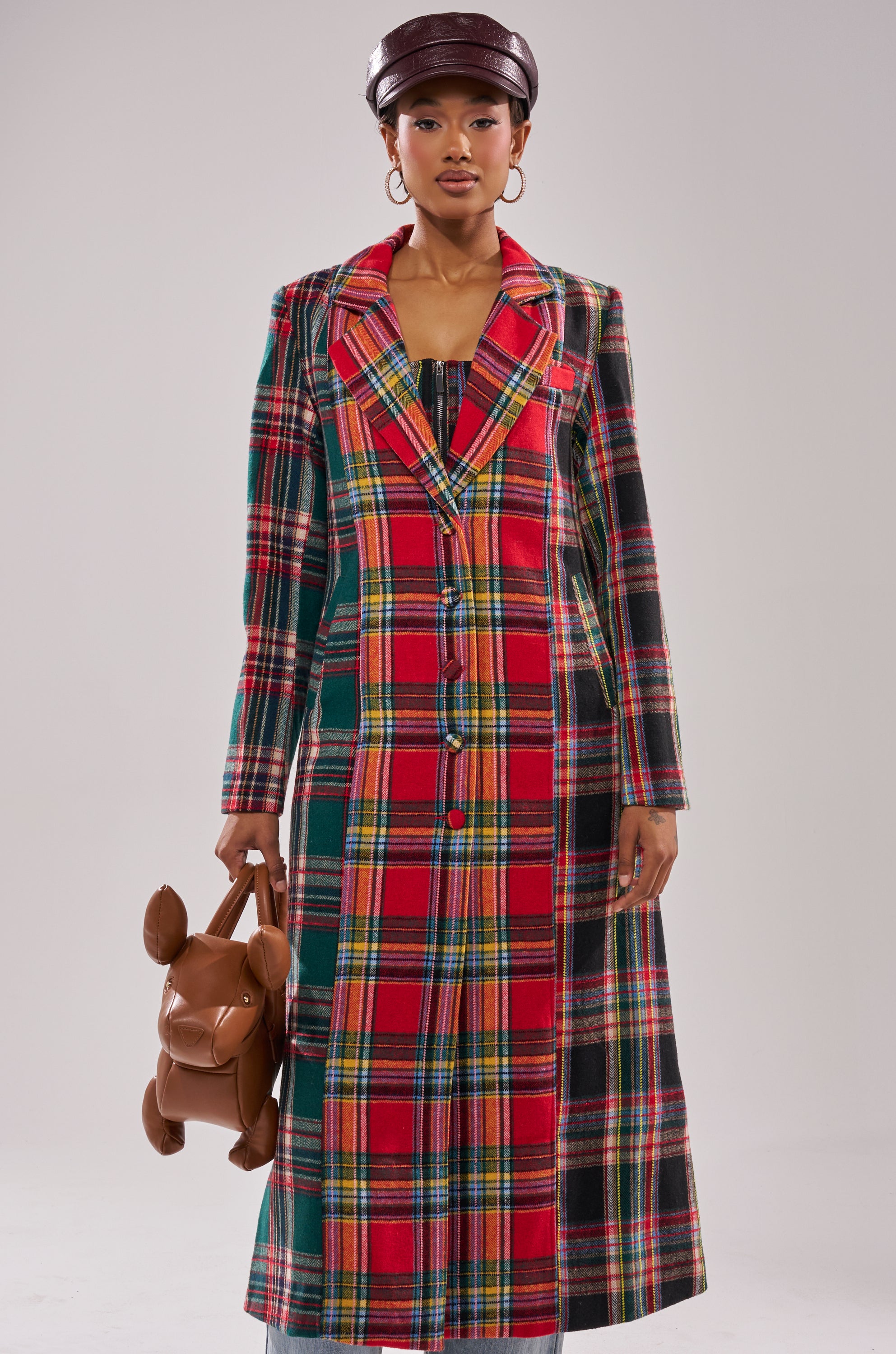 Wearing the TALK TO ME NICE PATCHWORK TRENCH and matching top, along with a brown leather cap and holding a brown dog-shaped handbag, a person stands against a plain light background.