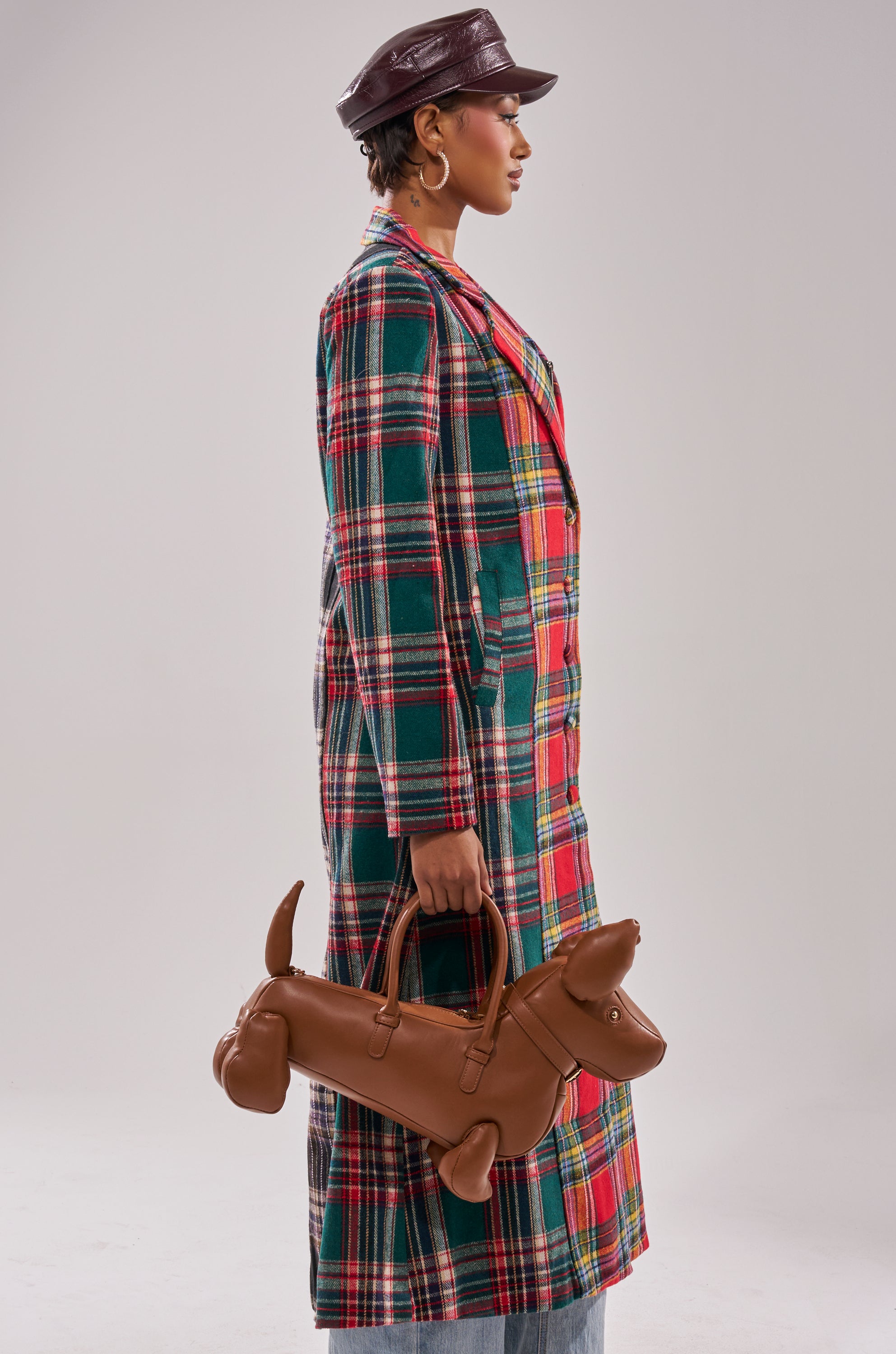 A person in profile wears the TALK TO ME NICE PATCHWORK TRENCH and a leather cap, holding a brown dachshund-shaped handbag. The background is plain and light-colored.