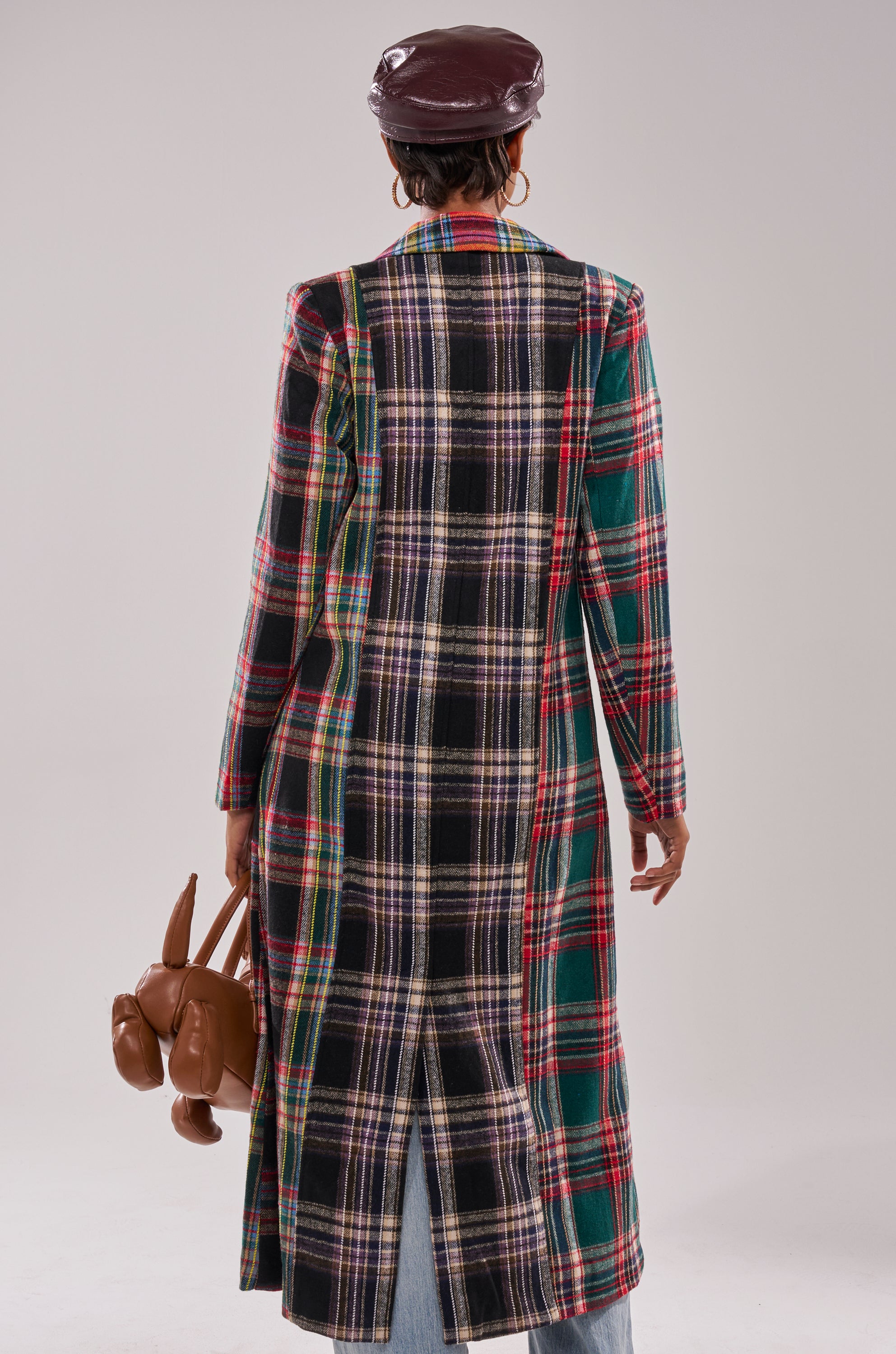 A person faces away wearing the AKIRA Label TALK TO ME NICE PATCHWORK TRENCH, light blue jeans, a maroon leather cap, and holding a brown dog-shaped handbag against a plain, light background.