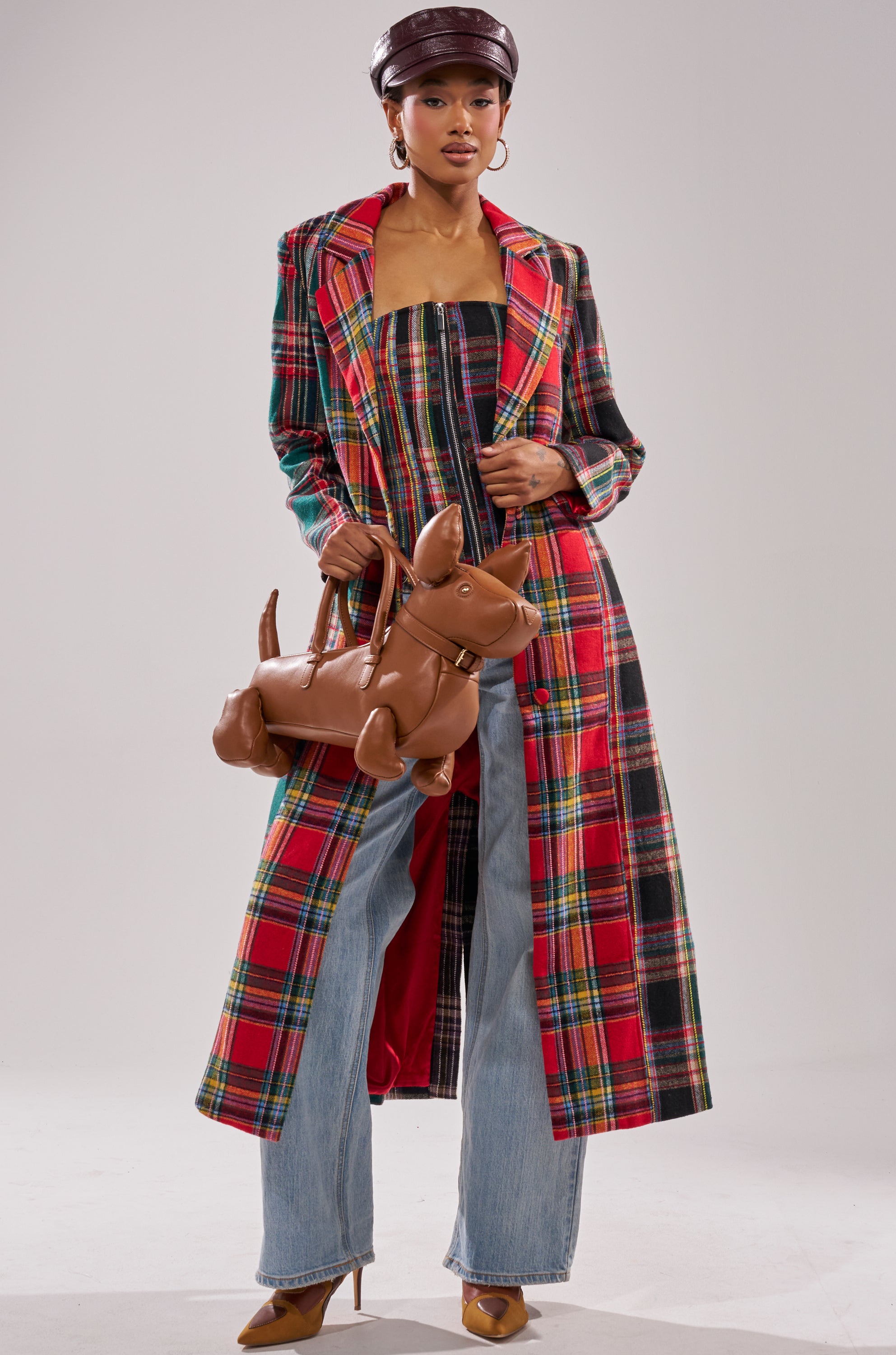 A woman in the AKIRA Label TALK TO ME NICE PATCHWORK TRENCH with a matching top, wide-leg jeans, tan heels, a brown newsboy cap, and a dog-shaped brown bag poses in a studio with a plain background.