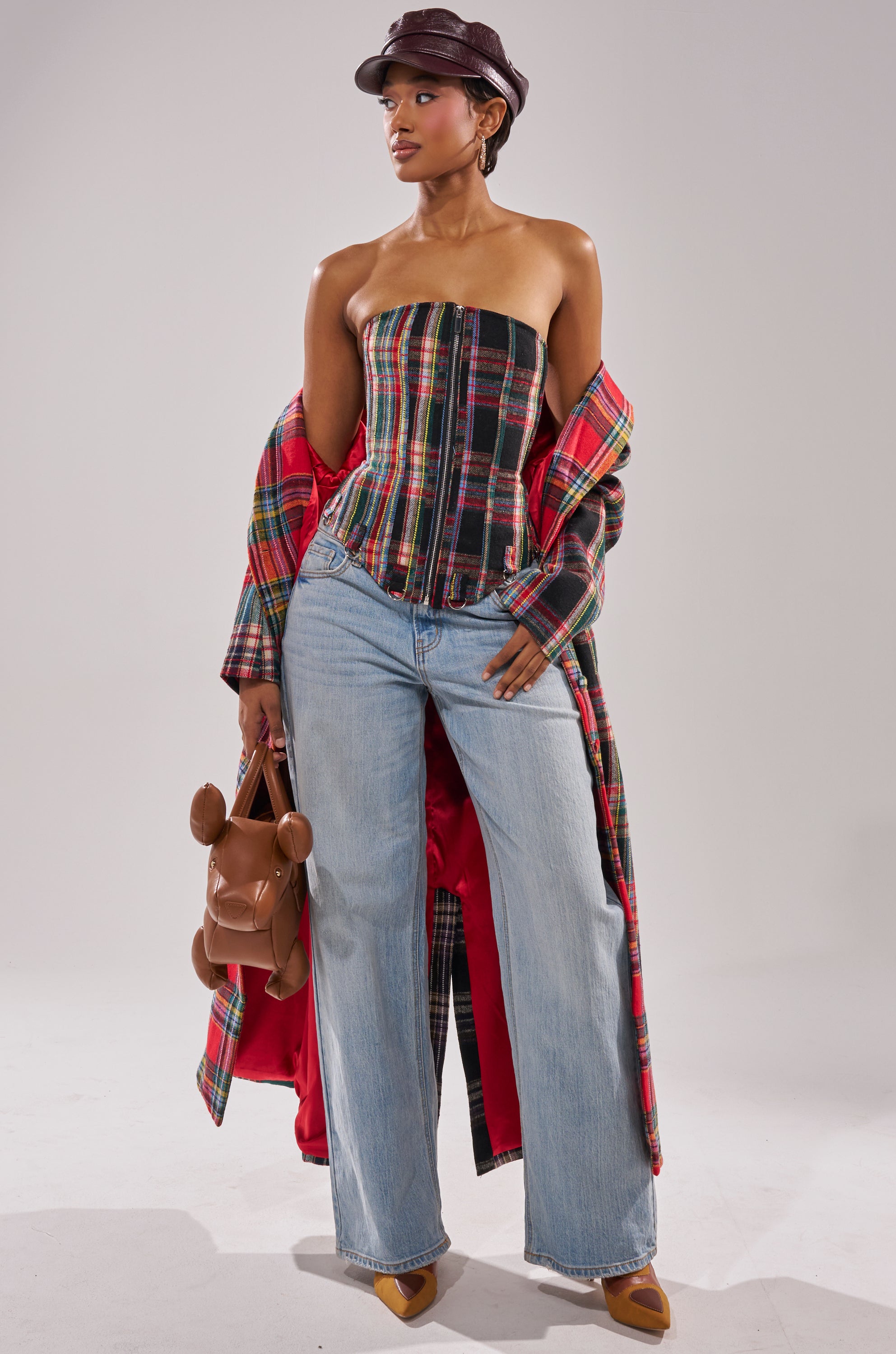 Wearing the TALK TO ME NICE PATCHWORK TRENCH, a woman pairs it with a corset top, wide-leg jeans, tan heels, and a brown cap while confidently holding a teddy bear-shaped purse against a plain background.