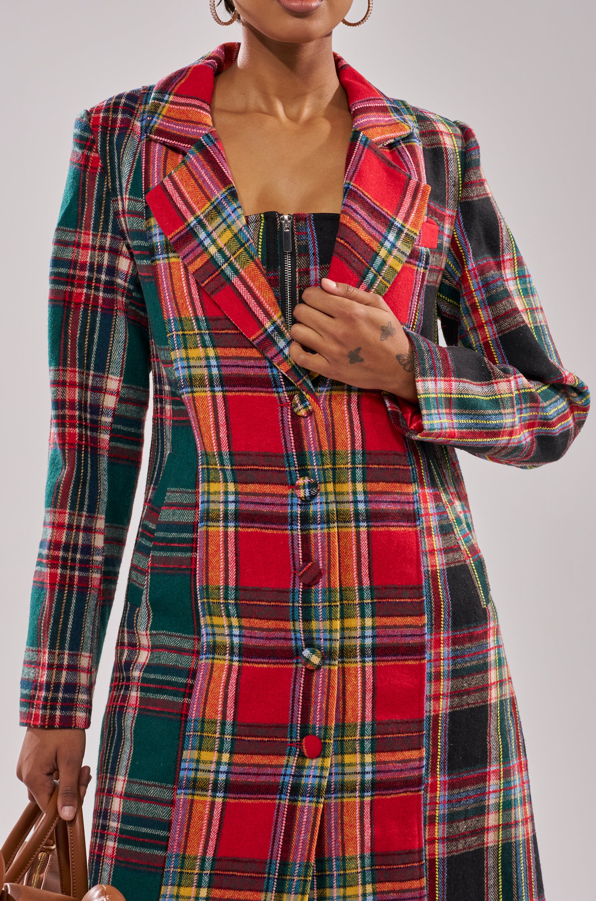 A person wearing the TALK TO ME NICE PATCHWORK TRENCH, a longline plaid coat with a notched collar and matching buttons, holds the lapel with one hand and carries a brown handbag in the other.