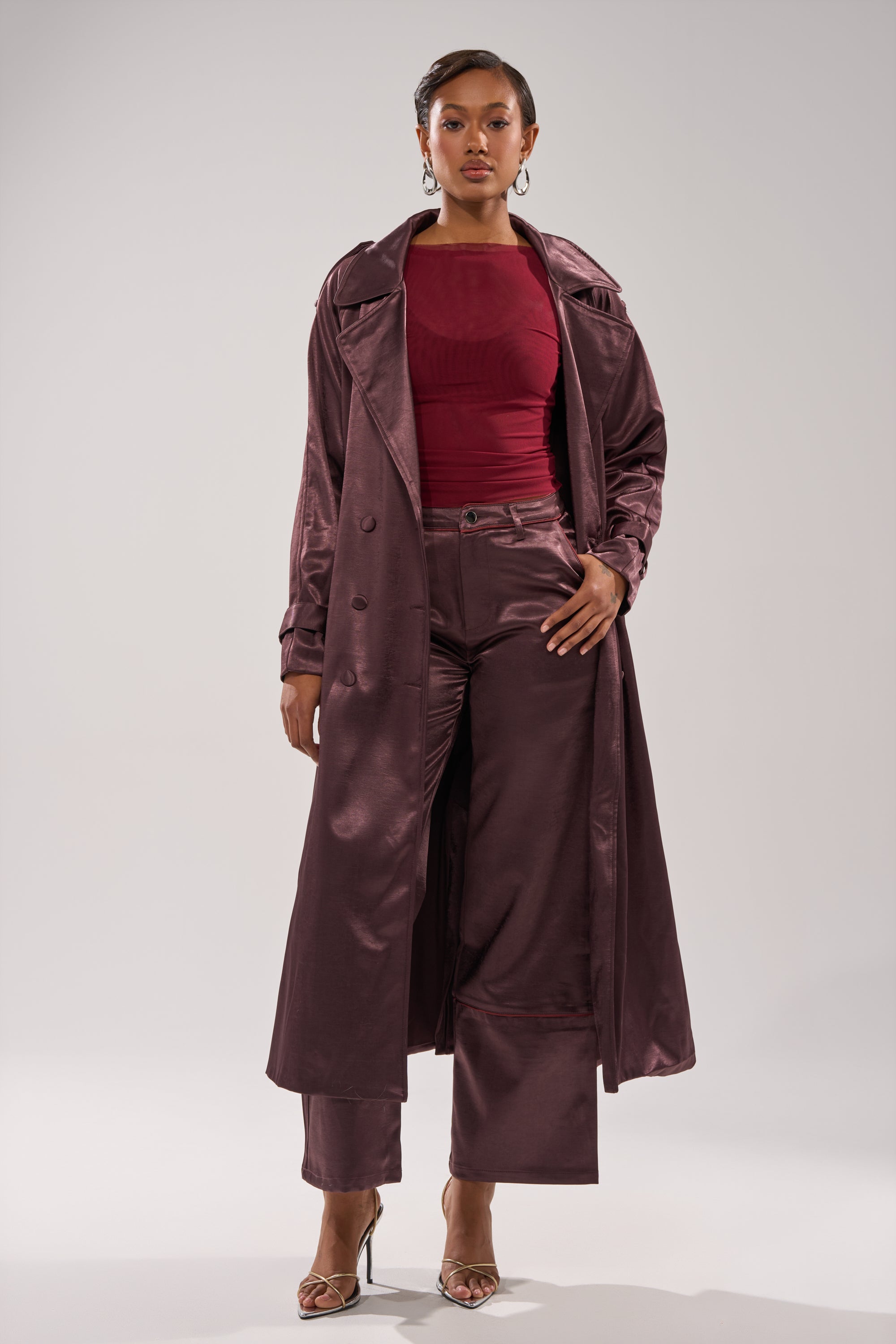 A woman stands confidently in the SLOW DOWN SATIN TROUSER, paired with a long mauve satin trench coat, a fitted burgundy top, and strappy high-heeled sandals against a plain light background. 0