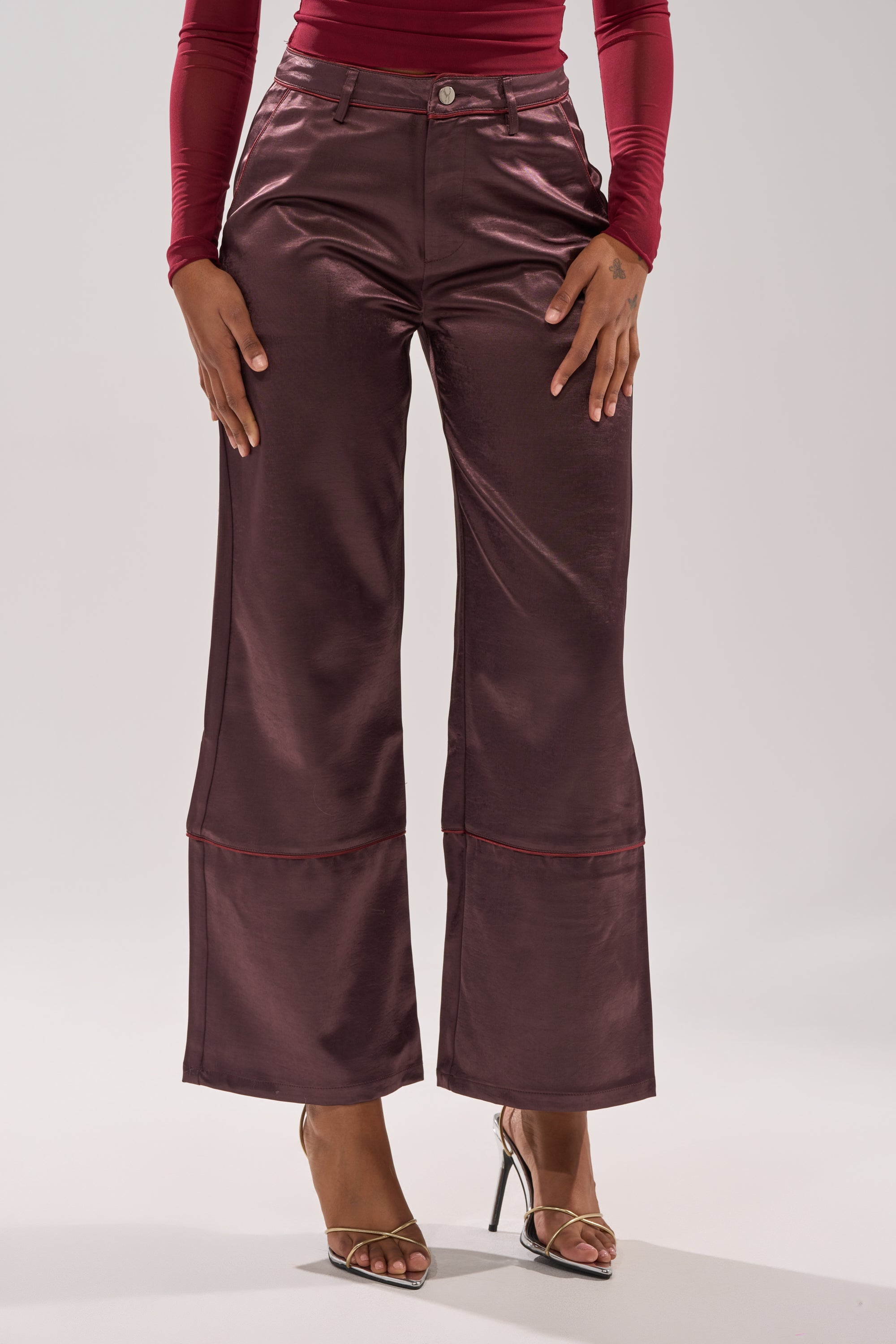 A person wears SLOW DOWN SATIN TROUSER—shiny burgundy wide-leg, high-rise pants—with a long-sleeve red top and strappy high-heeled sandals, standing against a plain light background. 1