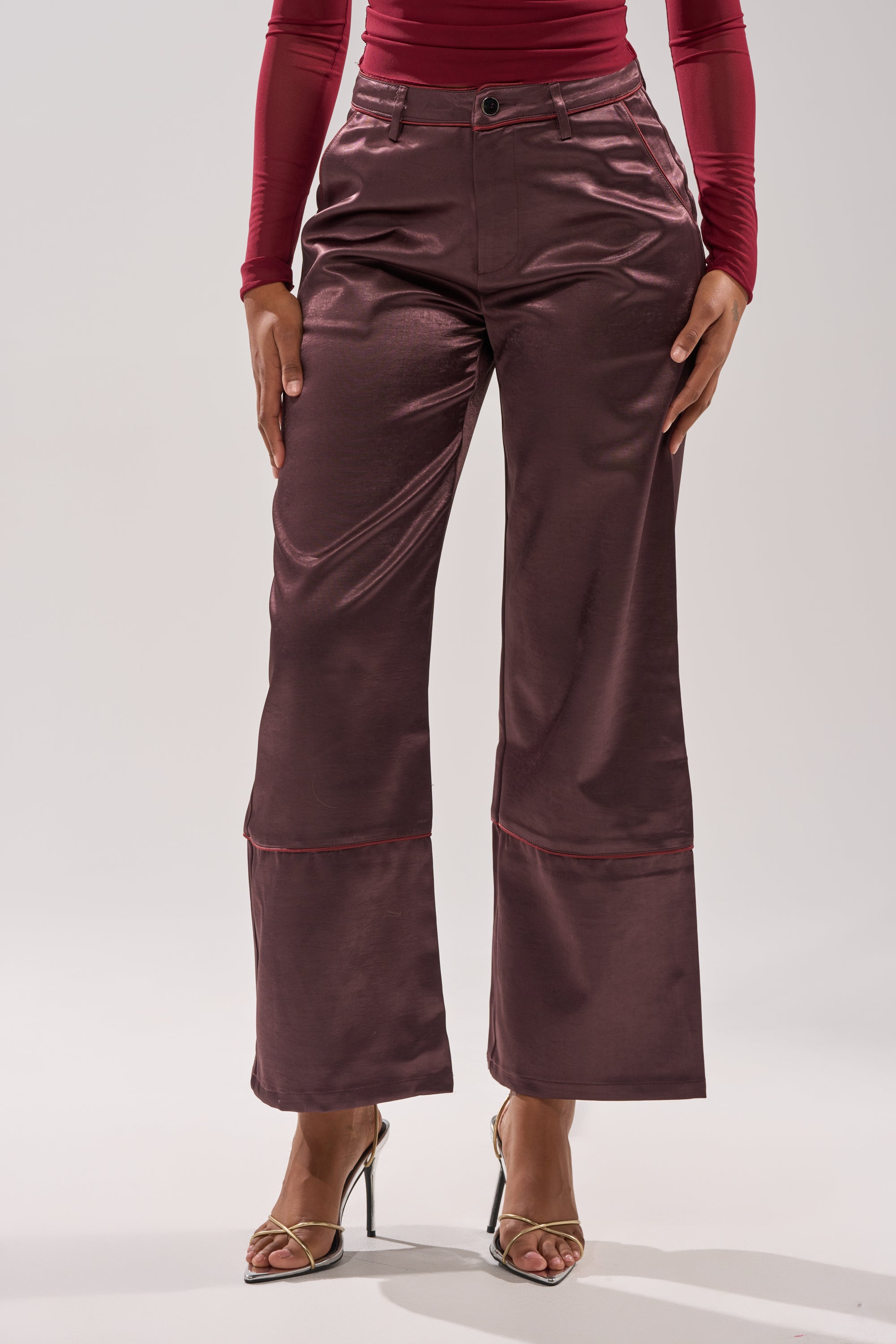 Someone stands against a plain light background wearing the SLOW DOWN SATIN TROUSER in shiny burgundy with a high-rise fit, paired with a matching long-sleeve top and open-toe high heel sandals. 2