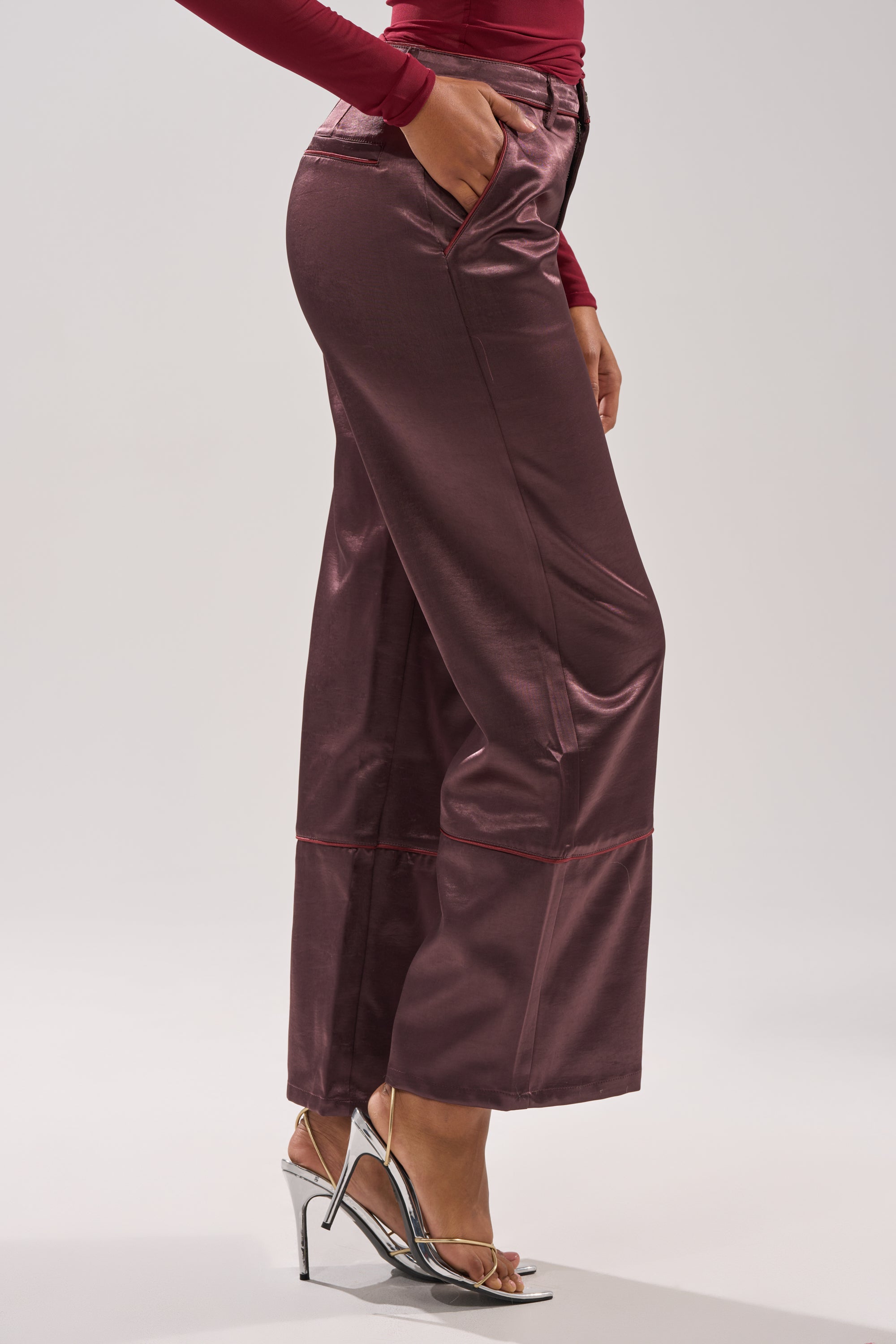 A person stands against a light background wearing the SLOW DOWN SATIN TROUSER—wide-leg, high-rise, shiny maroon satin pants—paired with a fitted long-sleeve top and silver high-heeled sandals. 3