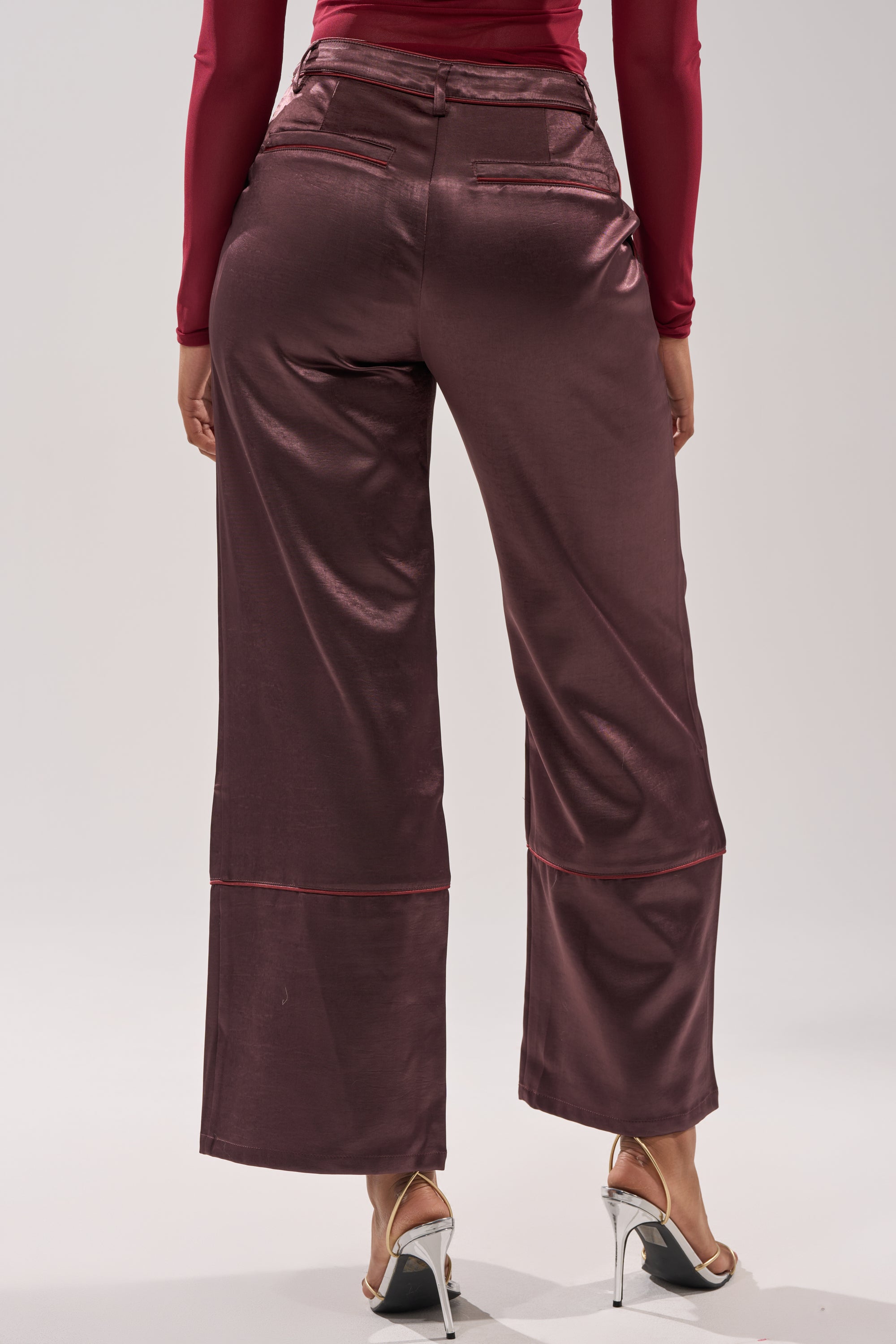 Wearing the SLOW DOWN SATIN TROUSER in shiny burgundy with a high-rise, wide-leg fit and a matching long-sleeve top, viewed from the back. Paired with silver high-heeled sandals. 4