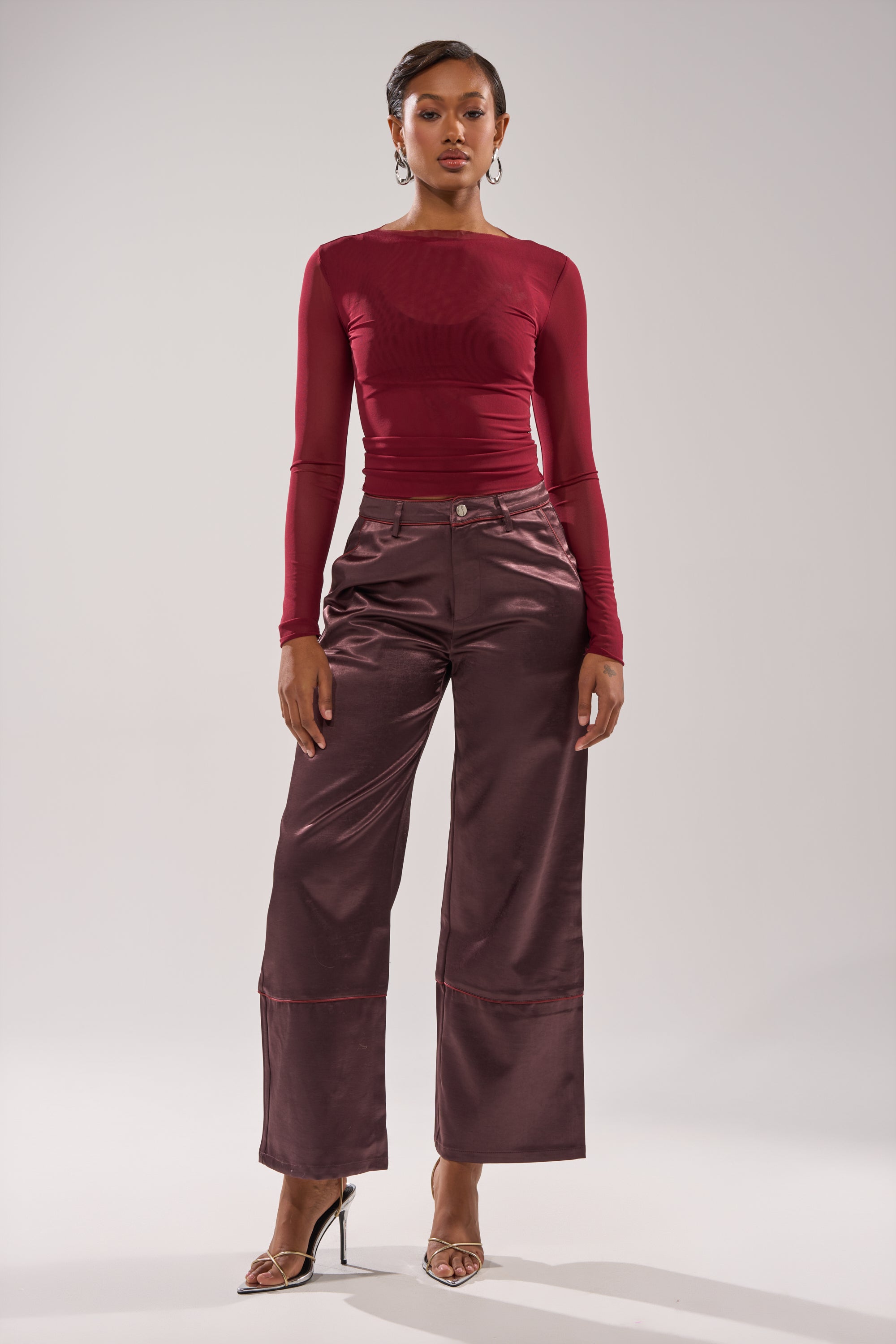 A woman faces forward in a fitted, long-sleeve burgundy top paired with the SLOW DOWN SATIN TROUSER—high-rise, wide-leg satin pants—and silver high-heeled sandals against a plain white background. 5
