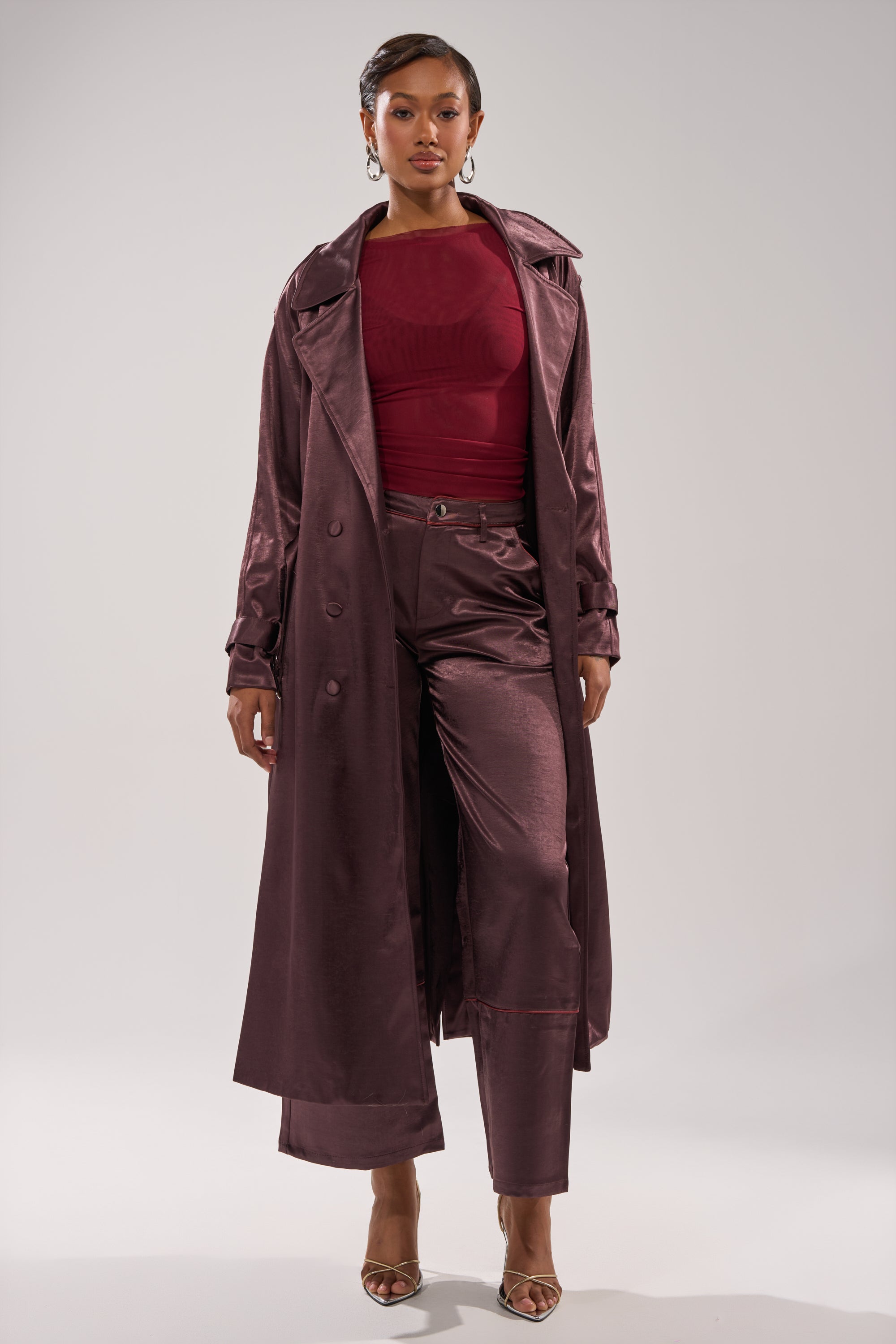 A woman stands confidently in SLOW DOWN SATIN TROUSER—high-rise, wide-leg satin pants—paired with a shiny burgundy trench coat, sheer red top, and gold heels against a plain light background. 6