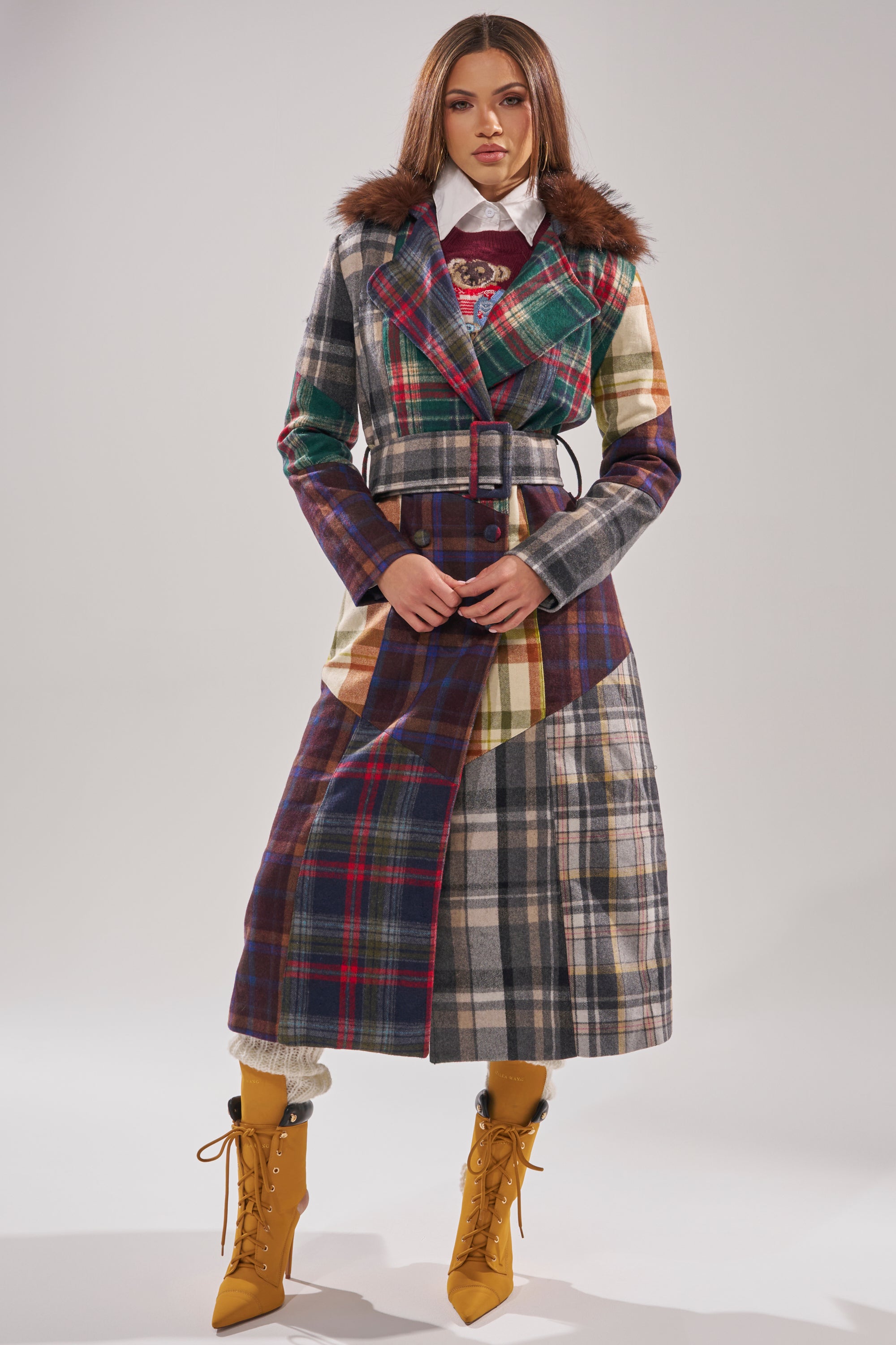 A woman stands against a plain background wearing the RILEY PATCHWORK TRENCH with a faux fur collar, paired with a white shirt, mustard yellow lace-up boots, and cream leg warmers. 0