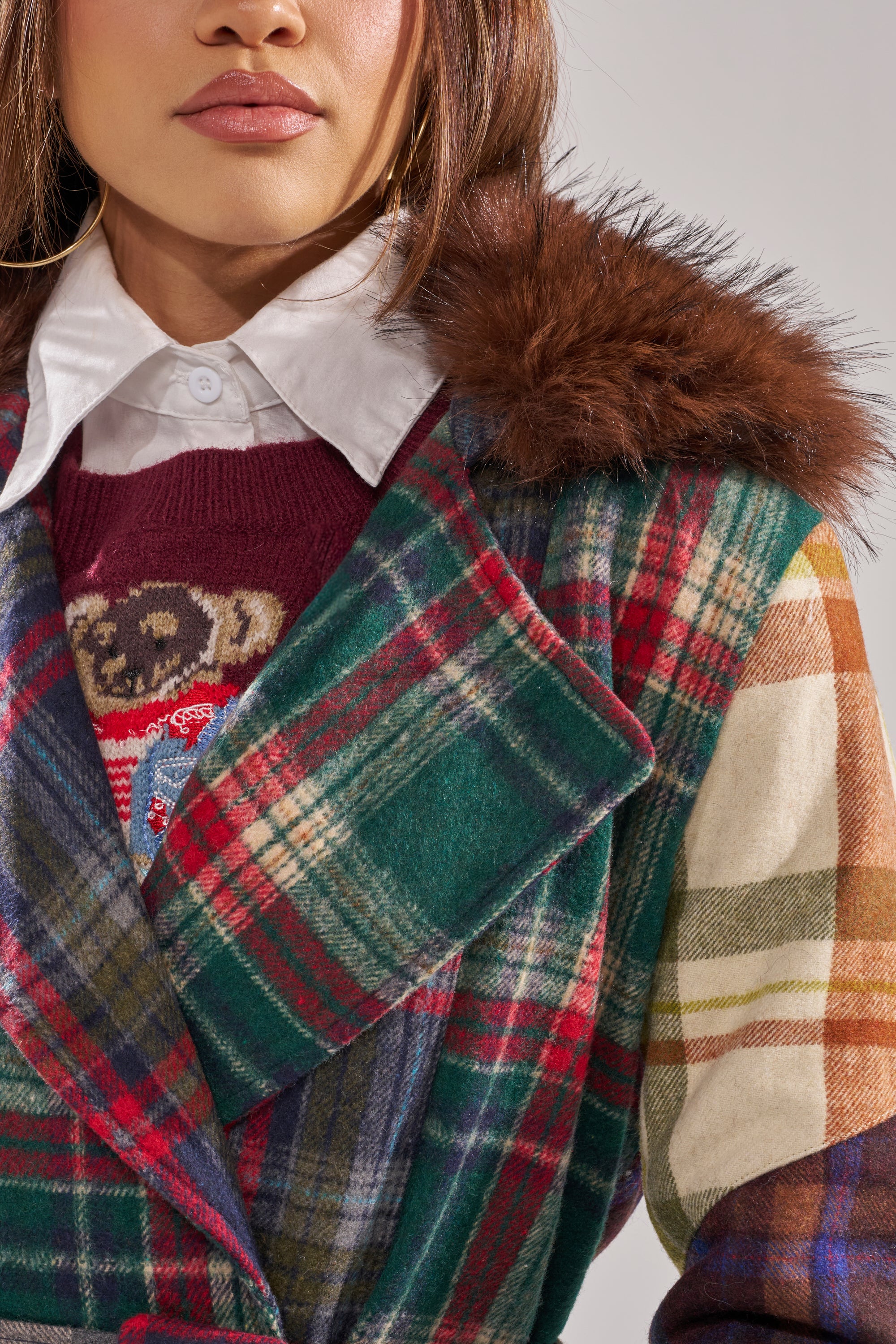 A woman wears the RILEY PATCHWORK TRENCH, a green and red plaid jacket with a brown faux fur collar, layered over a maroon teddy bear sweater and white collared shirt. The image shows her from shoulders to mid-chest. 9