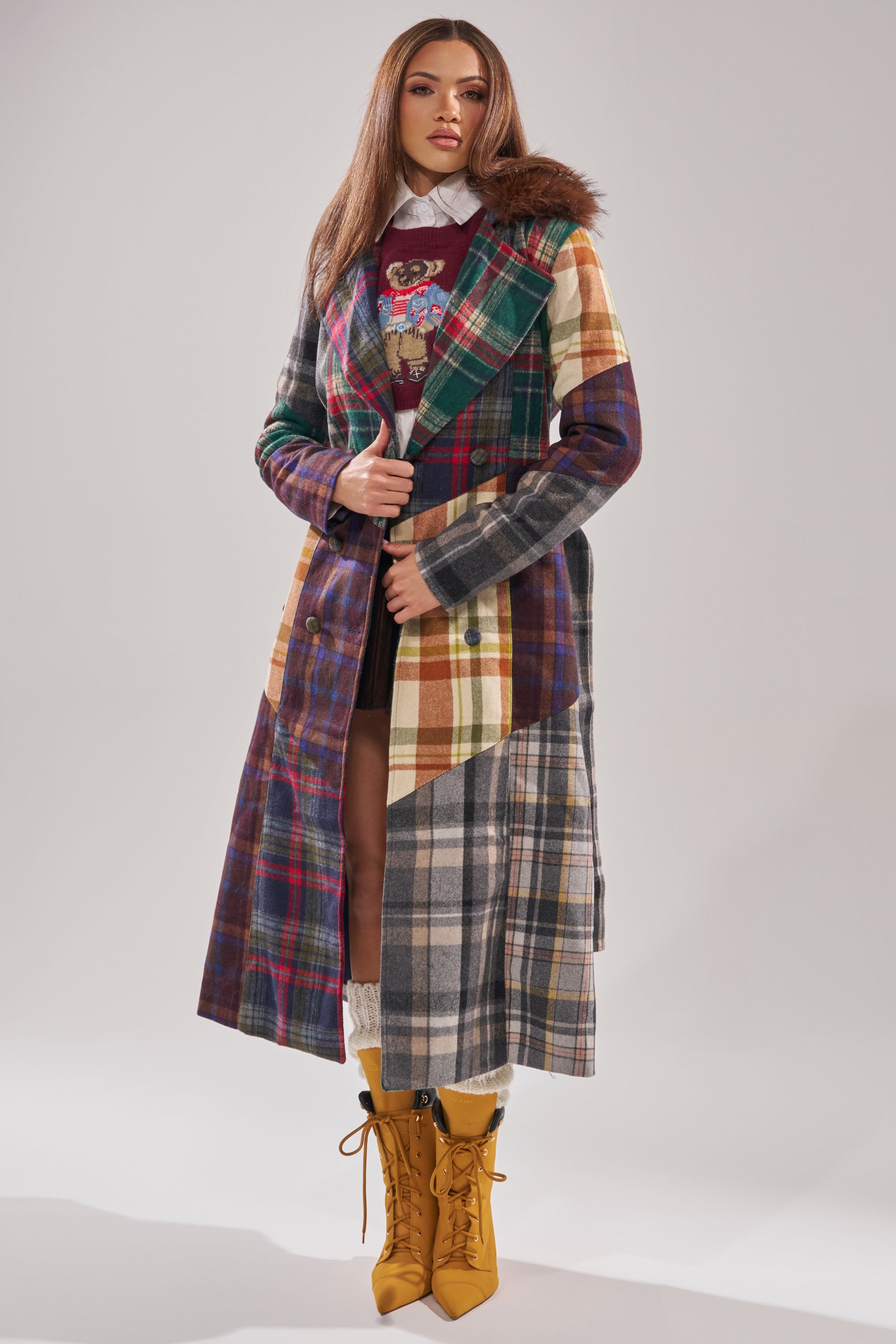 A woman models the RILEY PATCHWORK TRENCH—a plaid longline jacket with patchwork patterns and a faux fur collar—layered over a skirt and sweater, and styled with yellow lace-up boots against a plain light background. 1