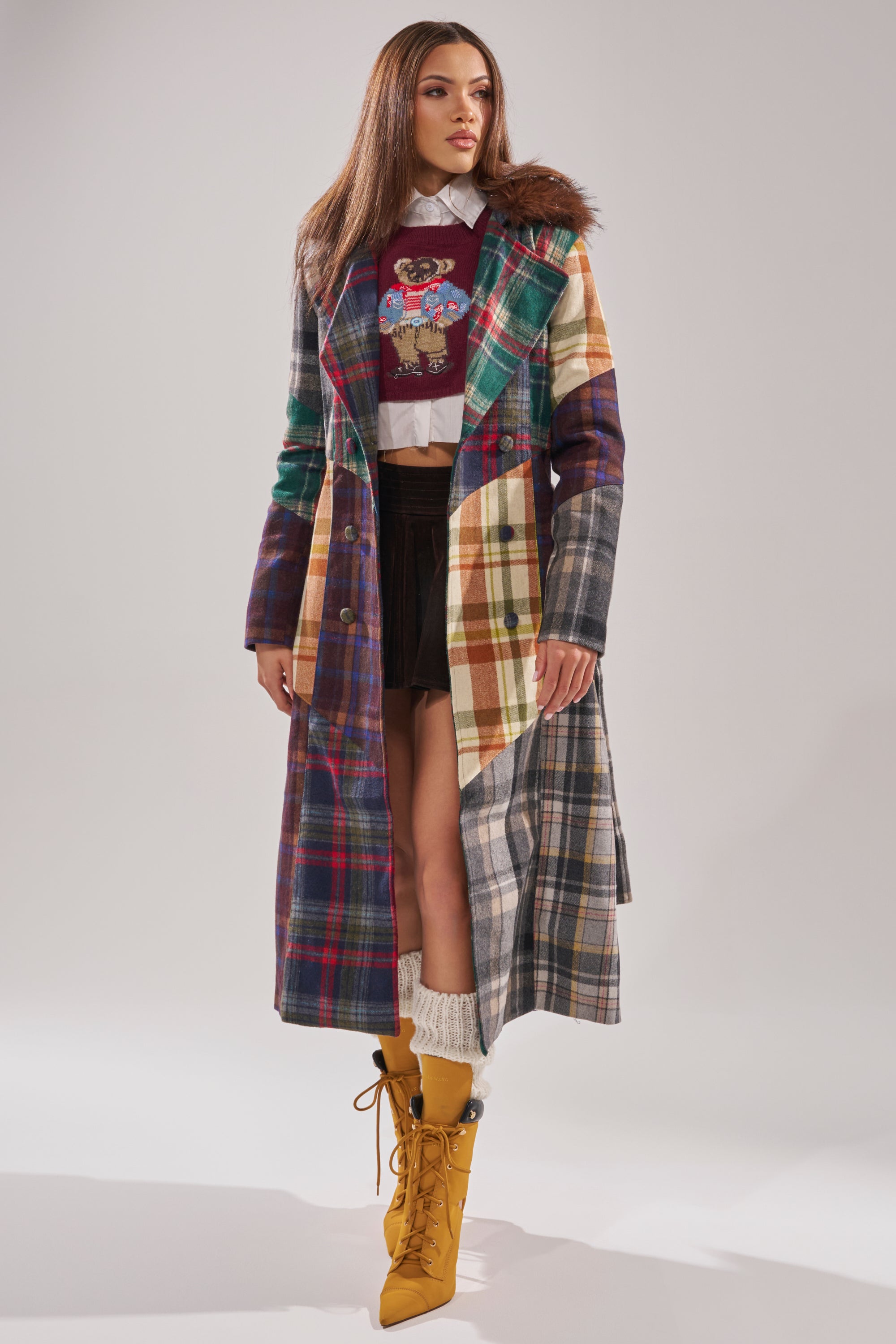 A woman poses against a plain background wearing the RILEY PATCHWORK TRENCH with a faux fur collar, styled over a graphic sweater, white shirt, black shorts, cream leg warmers, and yellow lace-up boots. 2