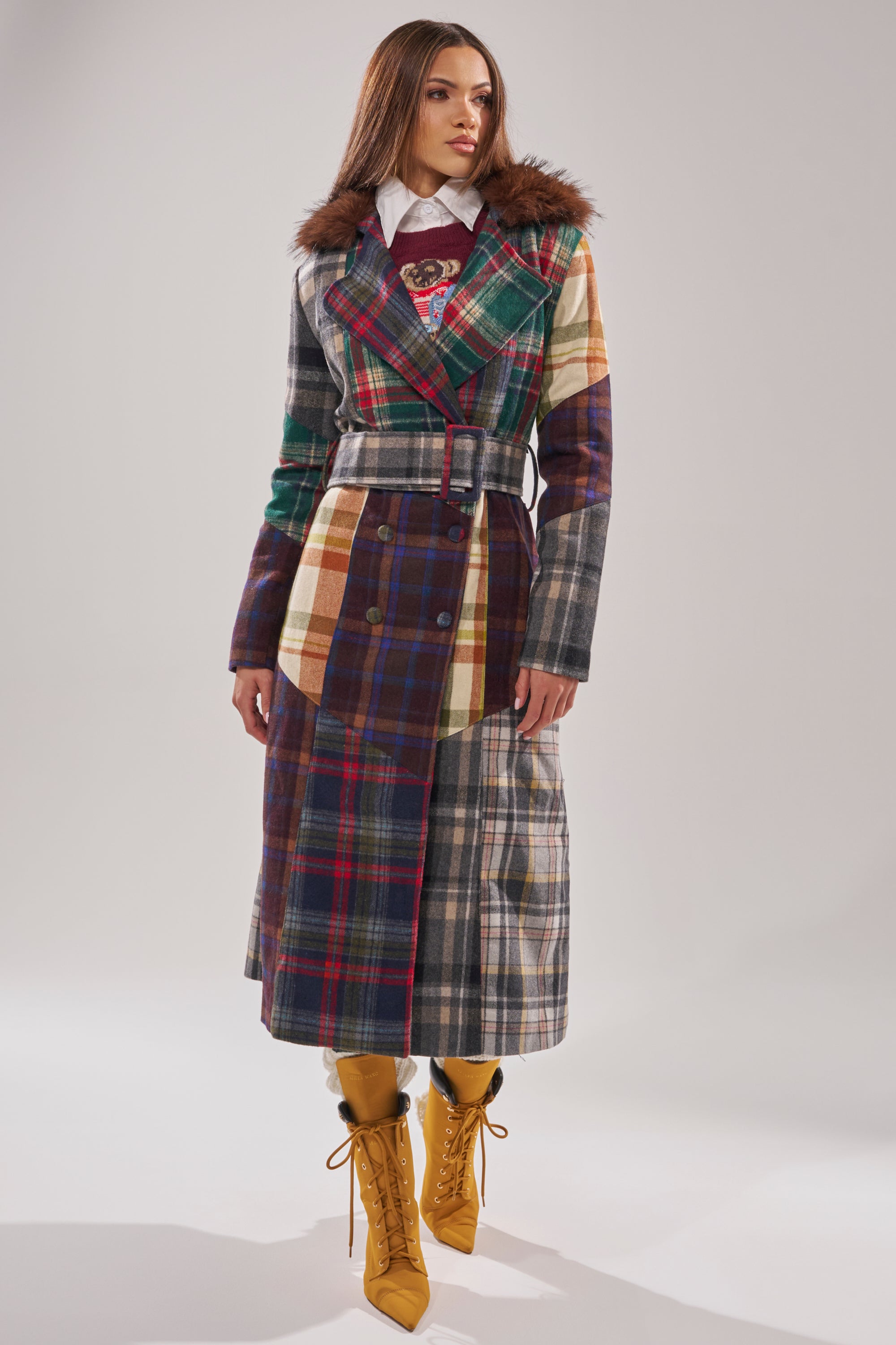 A woman stands facing forward against a plain light background, wearing the RILEY PATCHWORK TRENCH—a plaid longline jacket with a faux fur collar—paired with yellow lace-up boots and a white collared shirt. 5