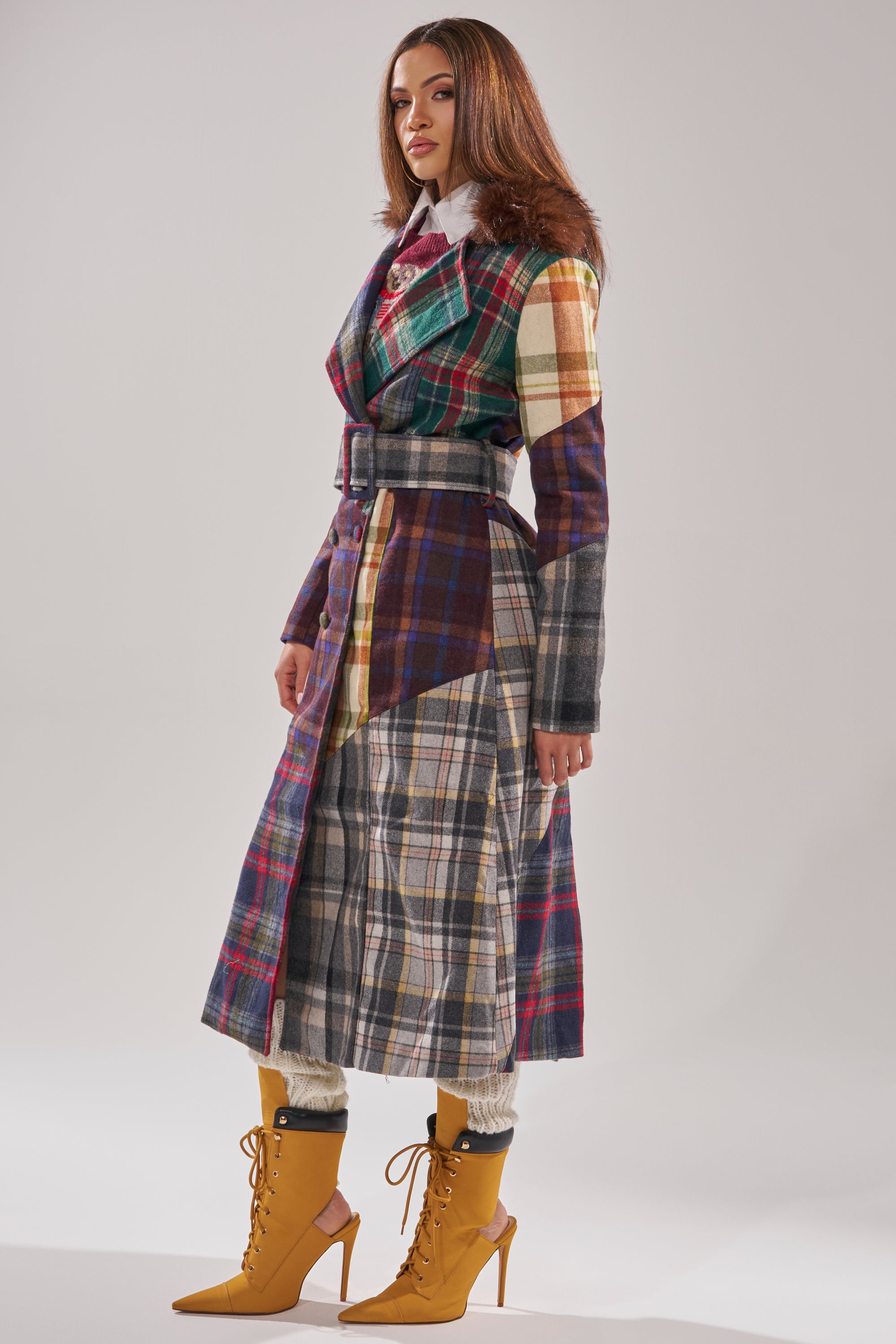 In a studio, a woman with long straight hair and a neutral expression models the RILEY PATCHWORK TRENCH, styled with white pants and mustard yellow lace-up high-heeled boots. 6