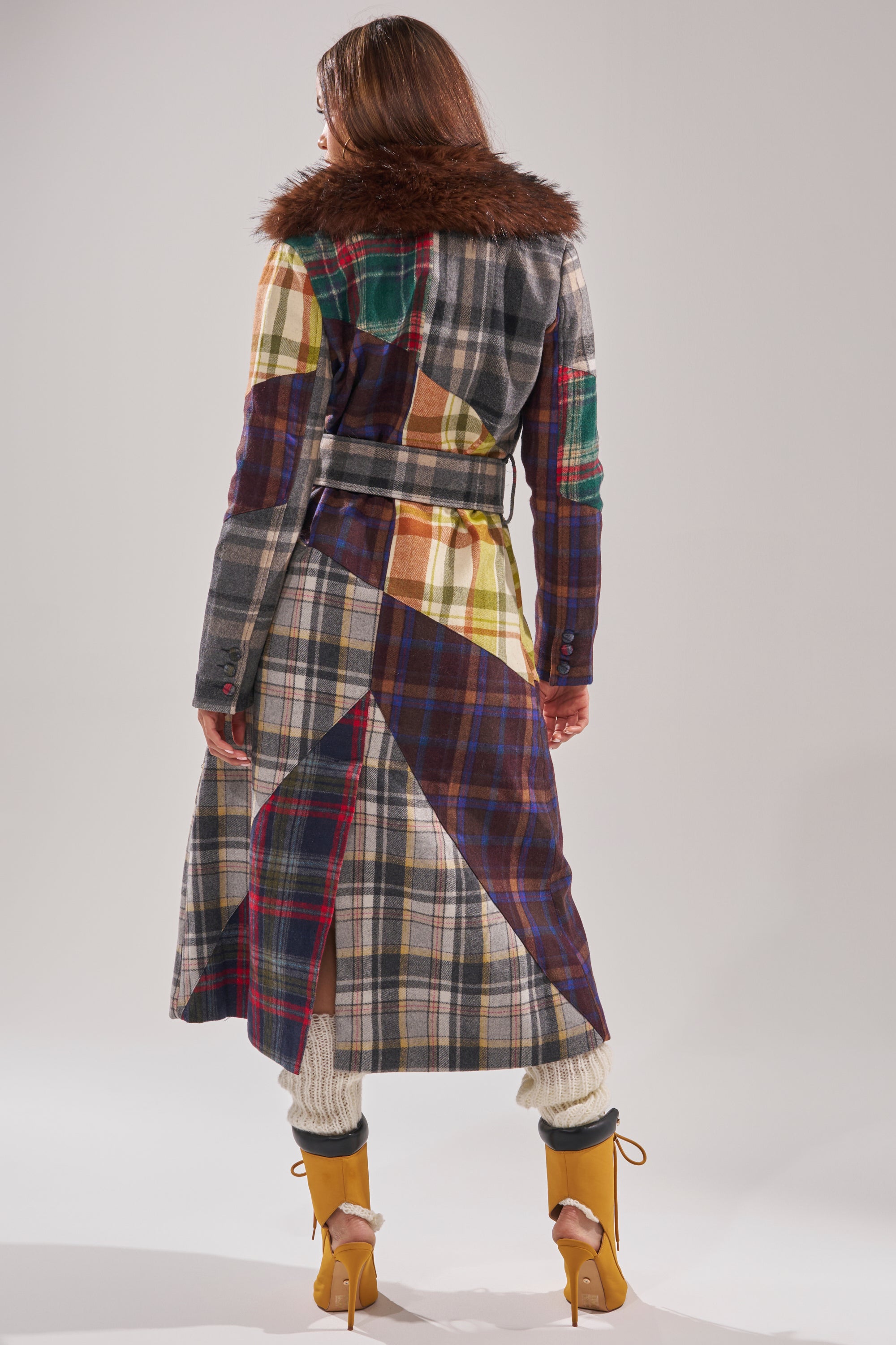 A person models the RILEY PATCHWORK TRENCH, a plaid longline jacket with a brown faux fur collar, paired with beige leg warmers and yellow high heels against a plain light background. 7