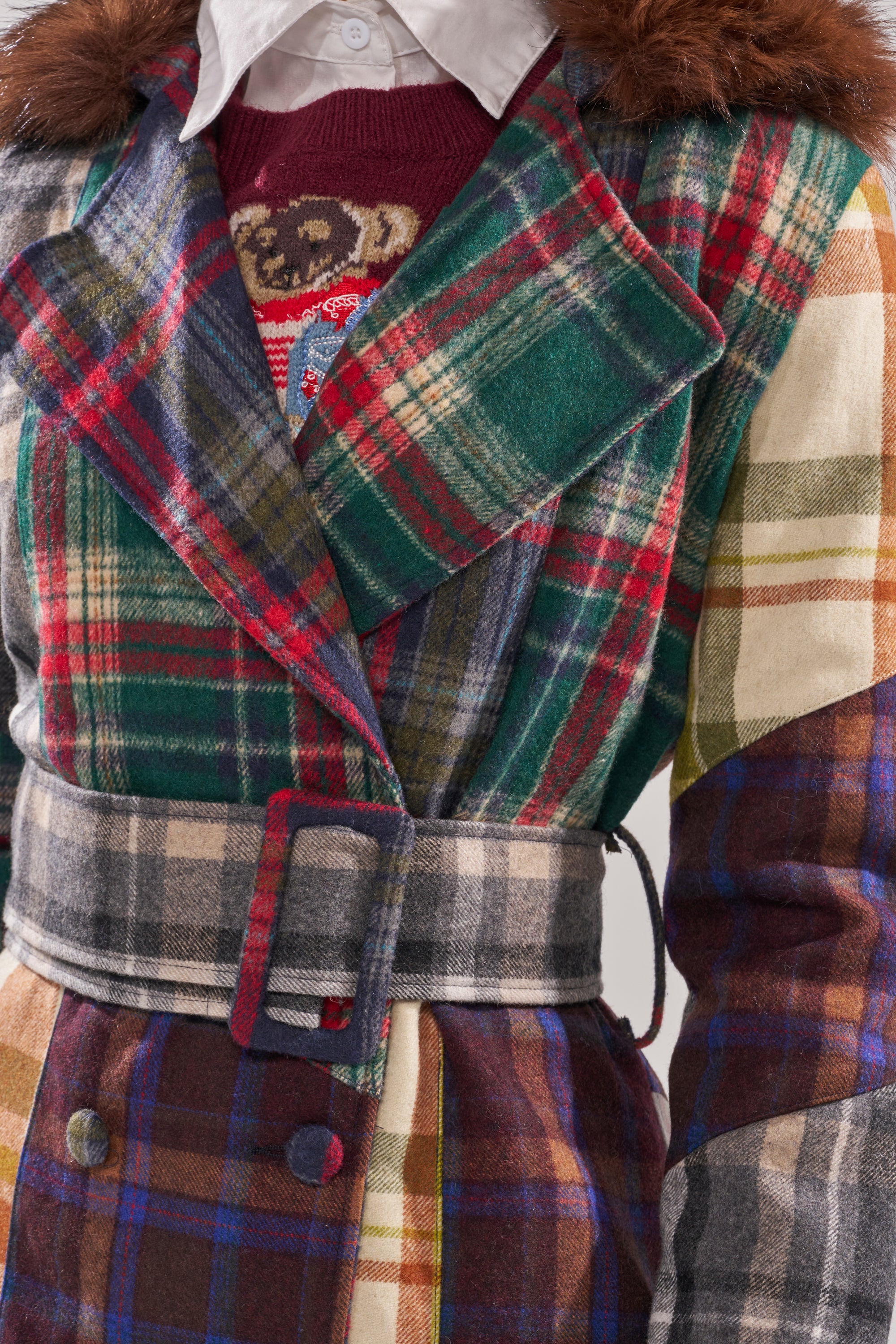 A close-up of a person wearing the RILEY PATCHWORK TRENCH with a belt, styled over a white collared shirt and a red sweater featuring an animal design. 8