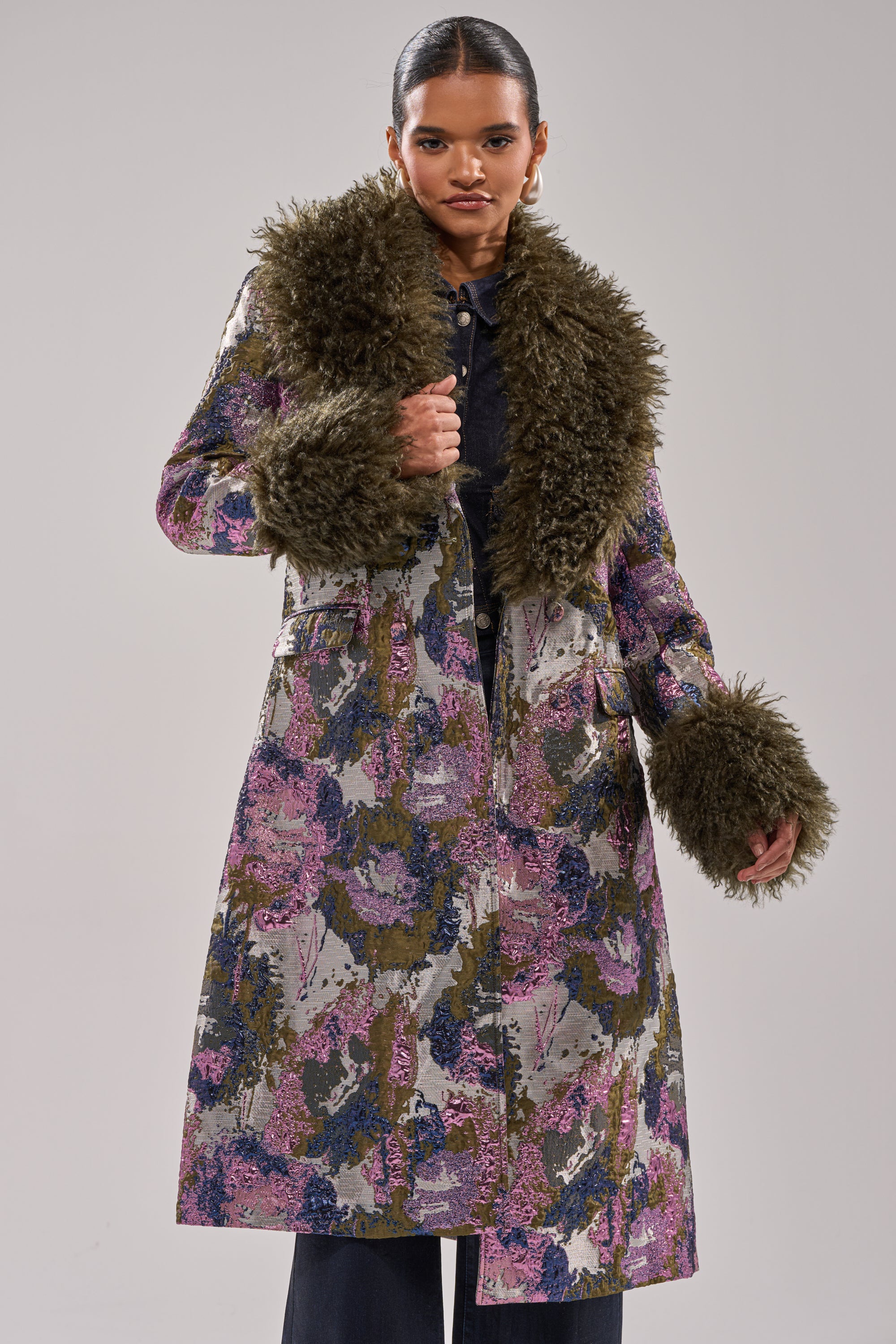 Wearing the GEORGINA BROCADE TRENCH, a long coat with floral metallic brocade and luxe faux fur trim on the collar and cuffs, a woman stands confidently against a plain background.