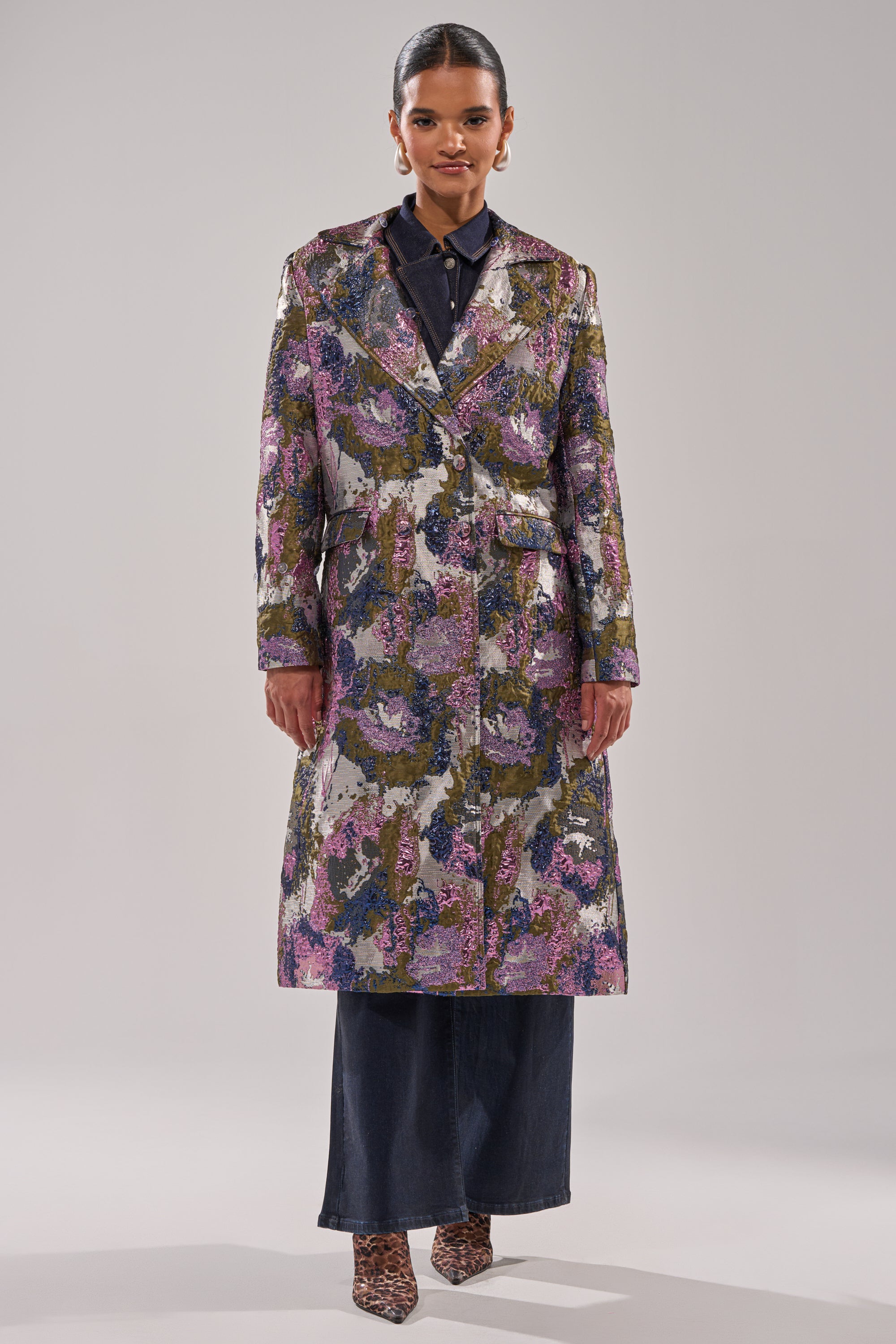 A woman faces forward wearing the GEORGINA BROCADE TRENCH in purple, green, and gold over a dark shirt and wide-leg pants. She has slicked-back hair, pointed shoes, and stands against a plain light background.