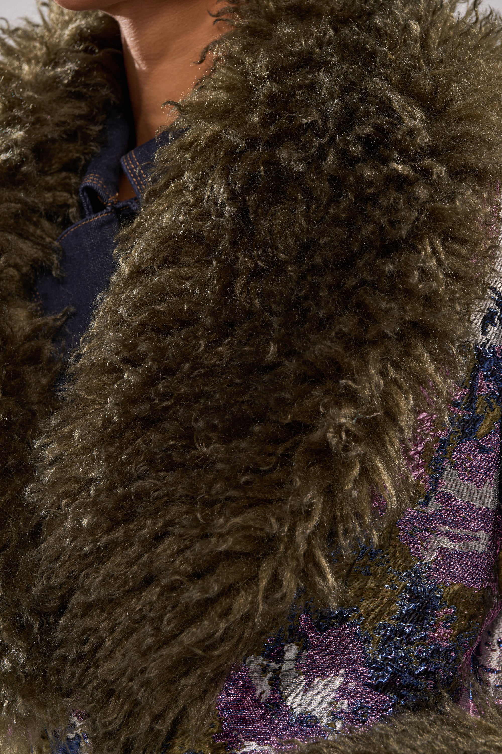 Close-up of a person wearing the GEORGINA BROCADE TRENCH, featuring a thick curly brown faux fur collar and colorful textured fabric with purple, blue, and metallic floral patterns.
