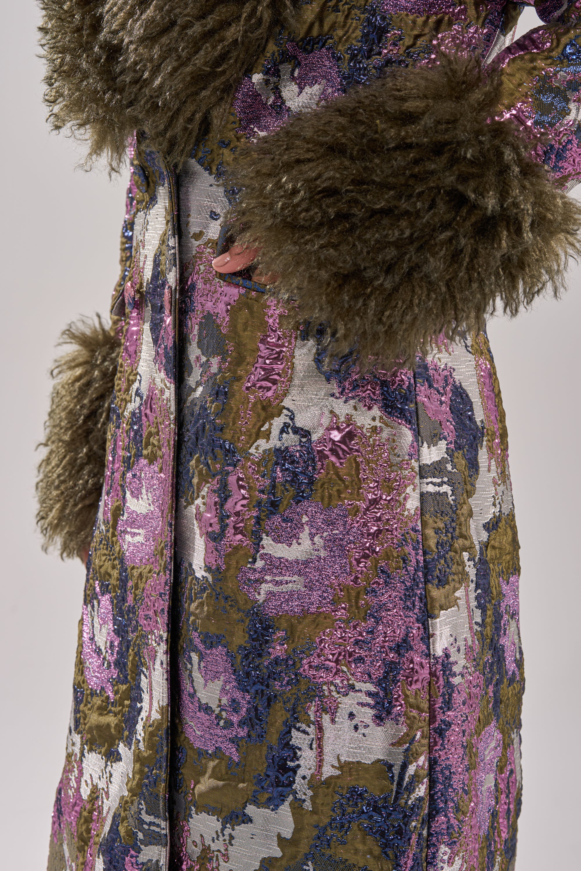 A person wearing the GEORGINA BROCADE TRENCH, featuring textured greenish-brown faux fur trim on the cuffs and collar, with a shiny abstract metallic design in purple, blue, and olive green; only the torso and part of an arm are shown.
