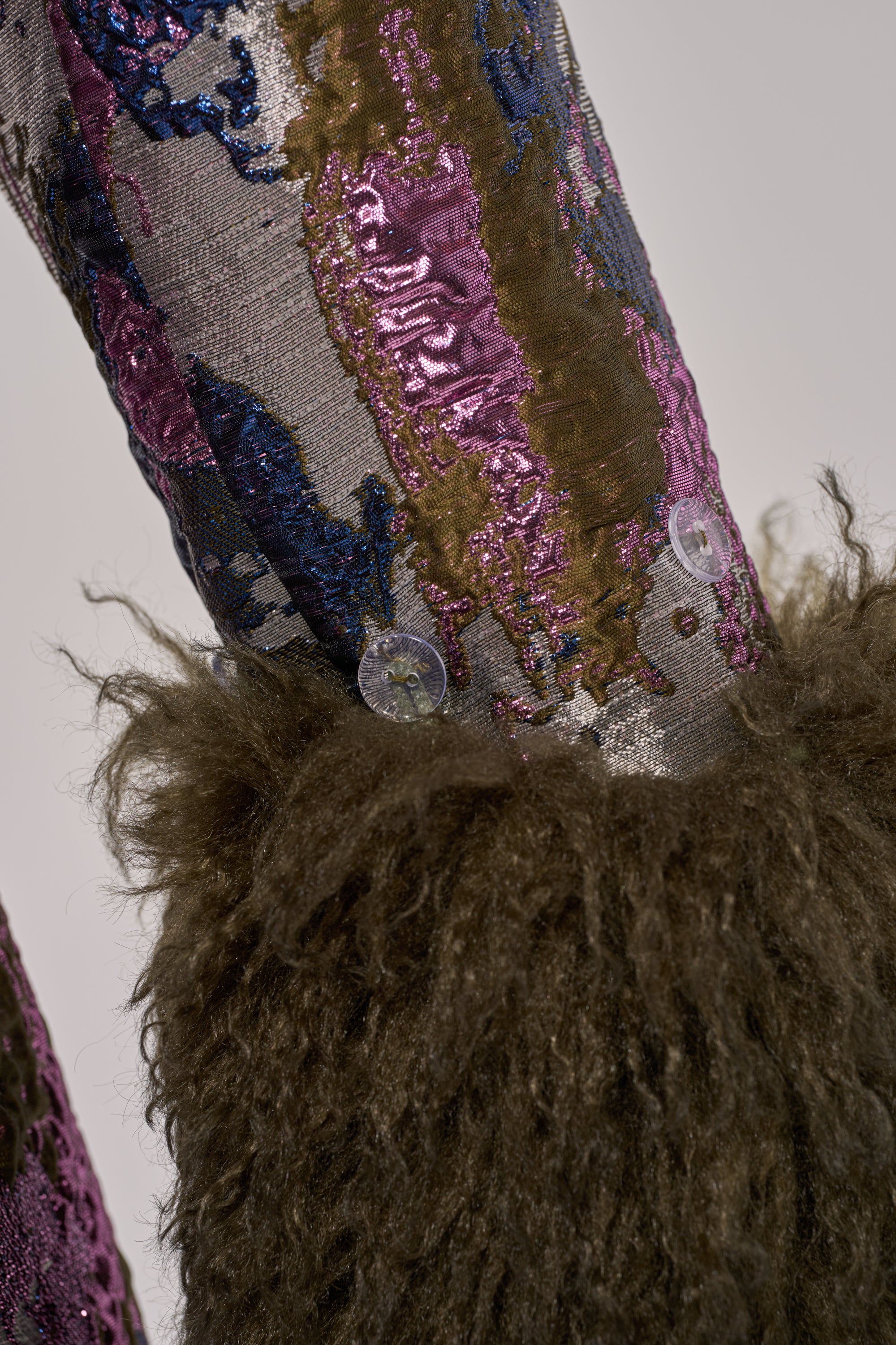 A close-up of the GEORGINA BROCADE TRENCH shows textured metallic fabric with shiny purple, blue, silver, and gold sequins, plus thick dark brown faux fur trim fastened by two transparent buttons.