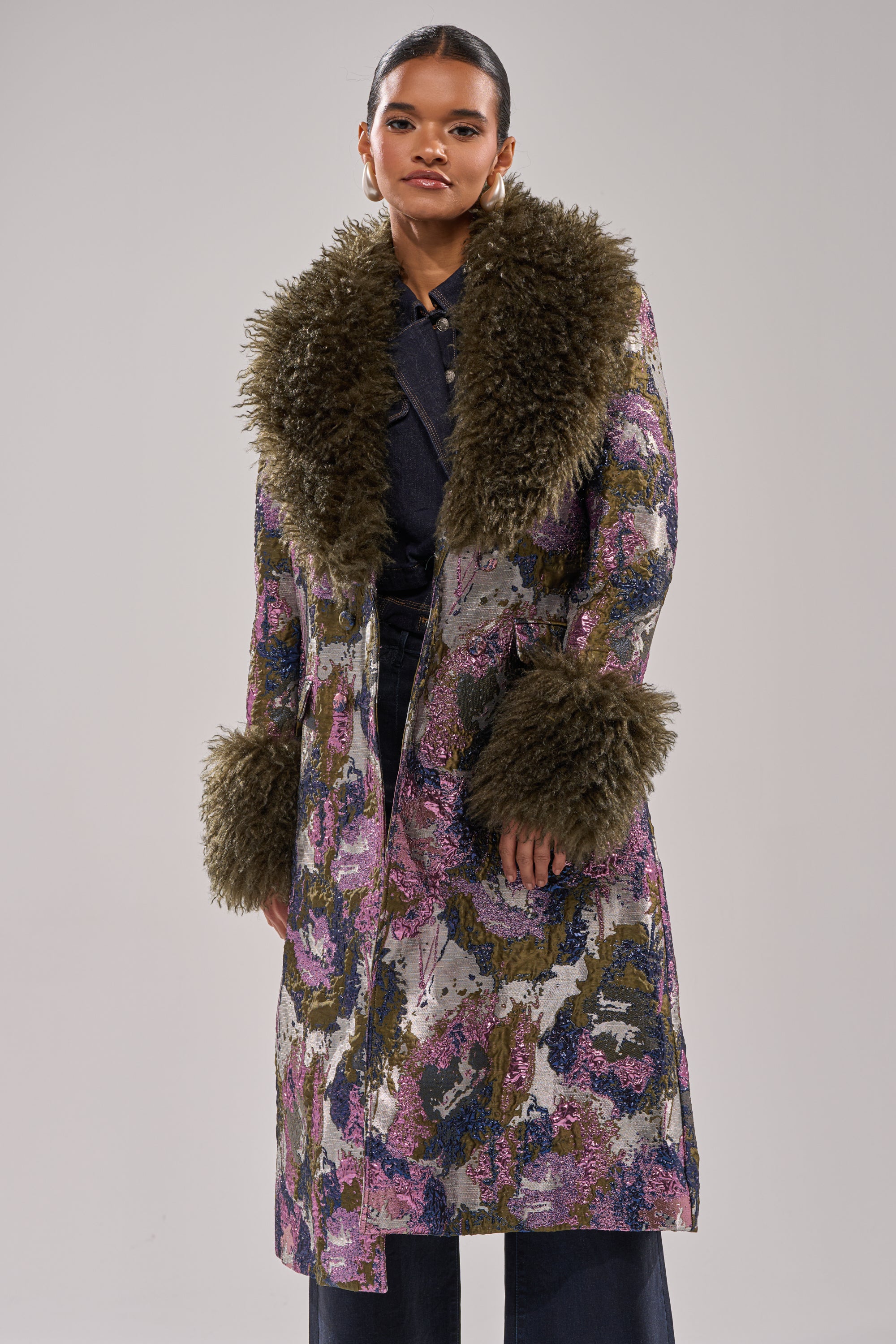 A woman wears the GEORGINA BROCADE TRENCH, a long metallic coat with a bold floral pattern and large, fluffy olive green faux fur trim on the collar and cuffs. She stands facing forward against a plain light gray background.