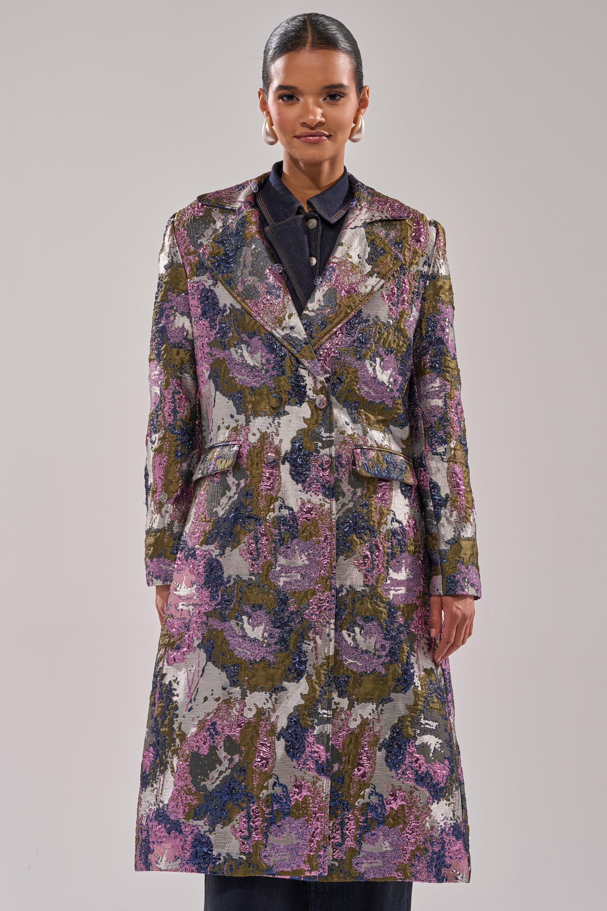 A woman faces forward in the GEORGINA BROCADE TRENCH—its purple, green, and silver metallic tones stand out over her dark shirt. With her hair pulled back and large hoop earrings, the plain background highlights her striking style.