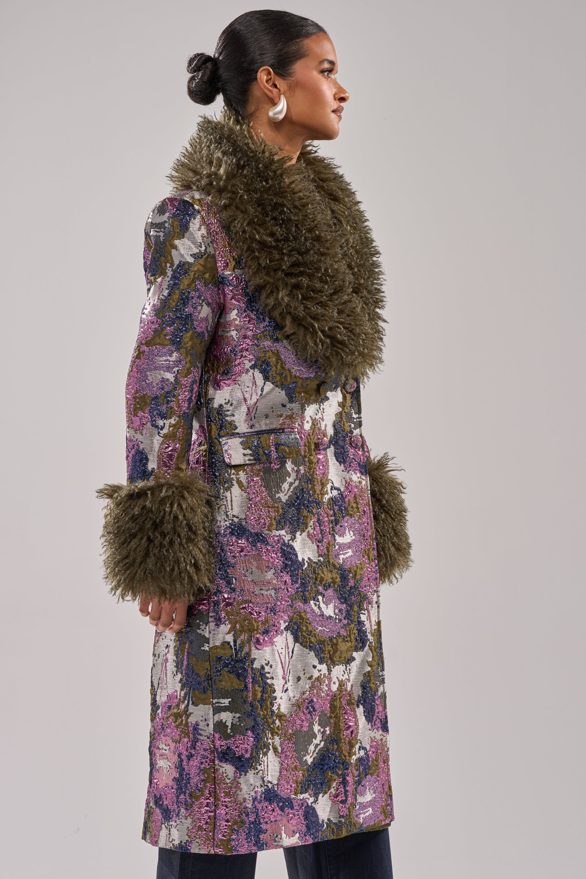 A woman stands sideways, wearing the GEORGINA BROCADE TRENCH—a long, colorful coat with a large faux fur collar and matching cuffs. Her dark hair is in a bun, and she completes the look with bold hoop earrings.