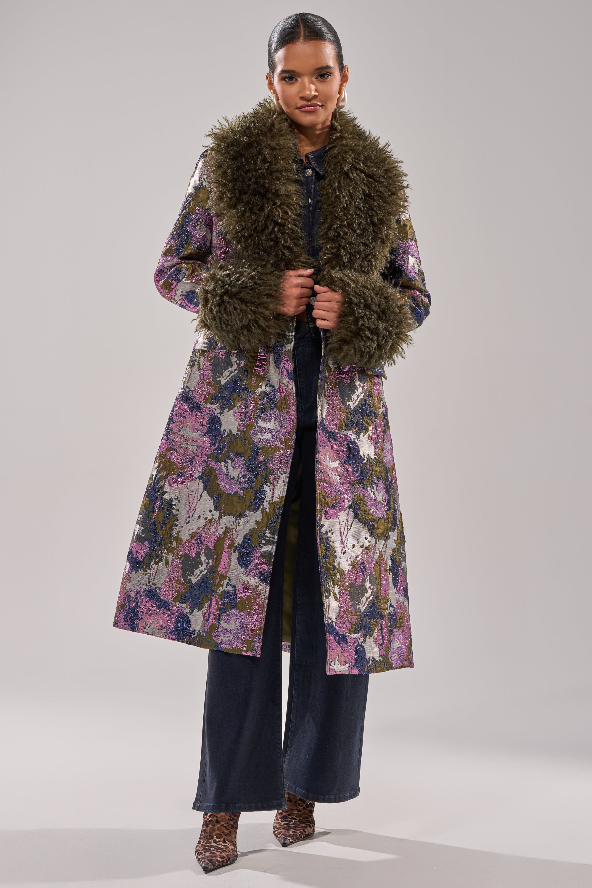 A woman poses against a plain light background wearing the GEORGINA BROCADE TRENCH, a metallic coat featuring a large dark brown faux fur trim on the collar and cuffs, styled with dark pants and heeled shoes.