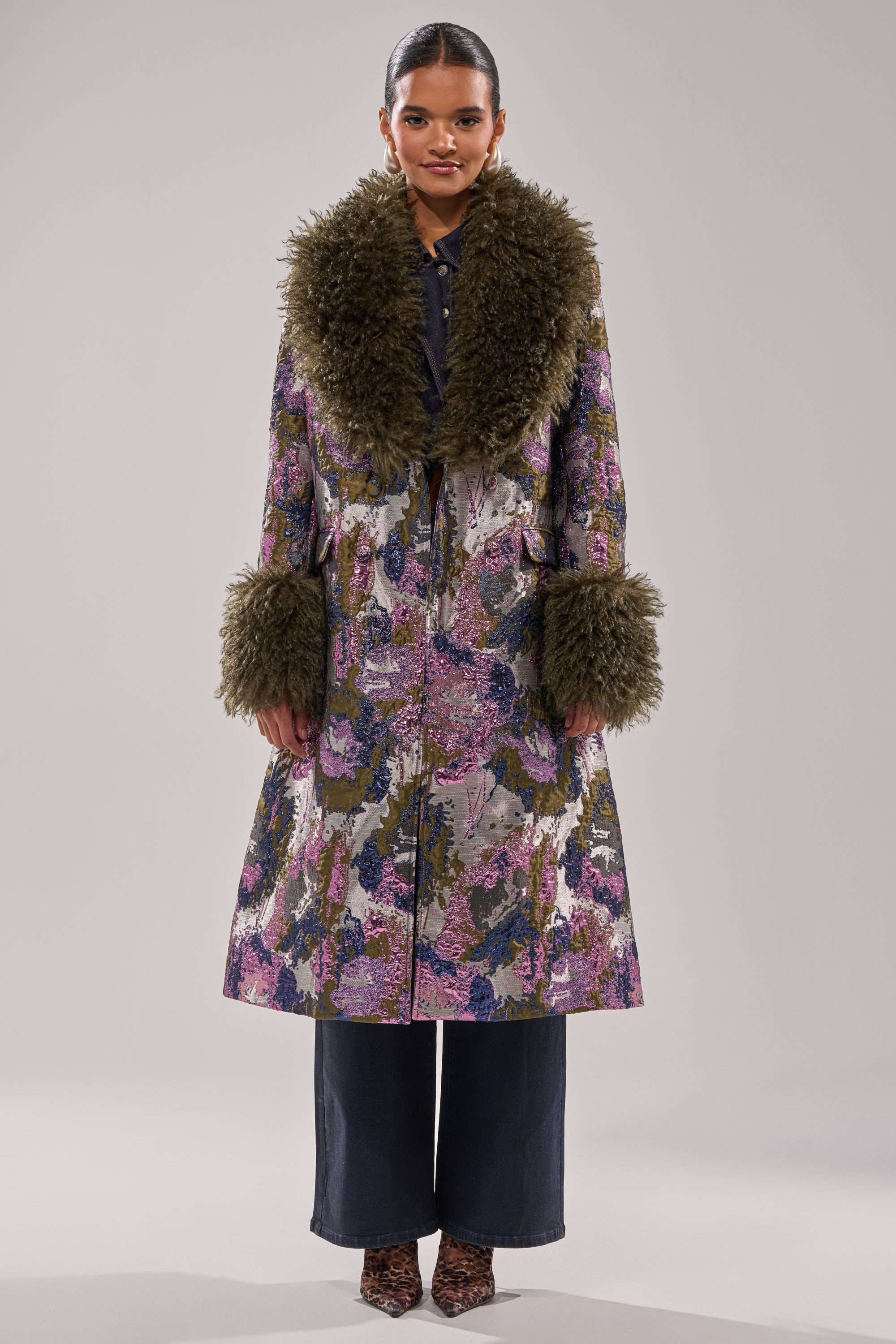 A woman wears the GEORGINA BROCADE TRENCH with large olive-green faux fur collar and cuffs, paired with dark wide-leg pants and animal print shoes, standing against a plain light background.
