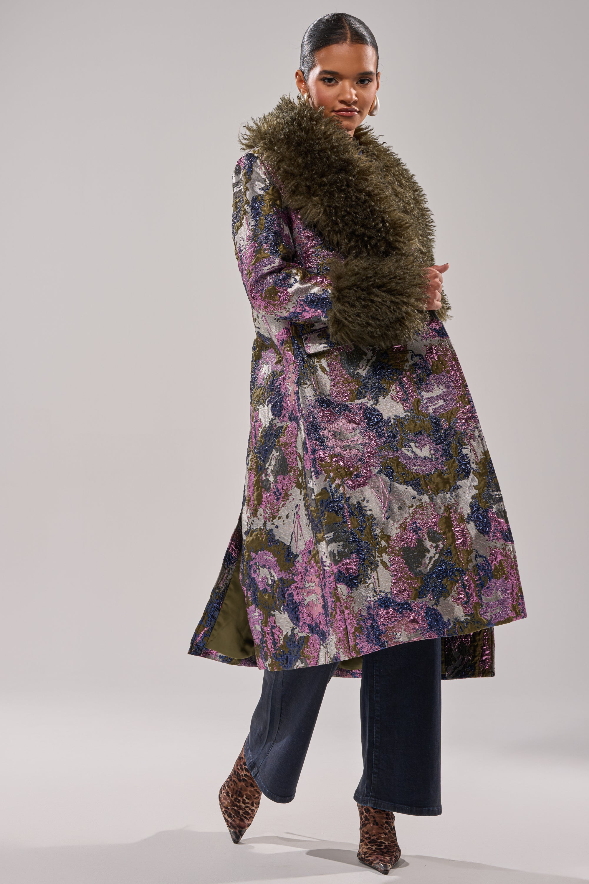 A woman showcases the GEORGINA BROCADE TRENCH—a long purple and green patterned coat with large faux fur collar and cuff trims—paired with dark pants and animal-print heeled boots against a light background.