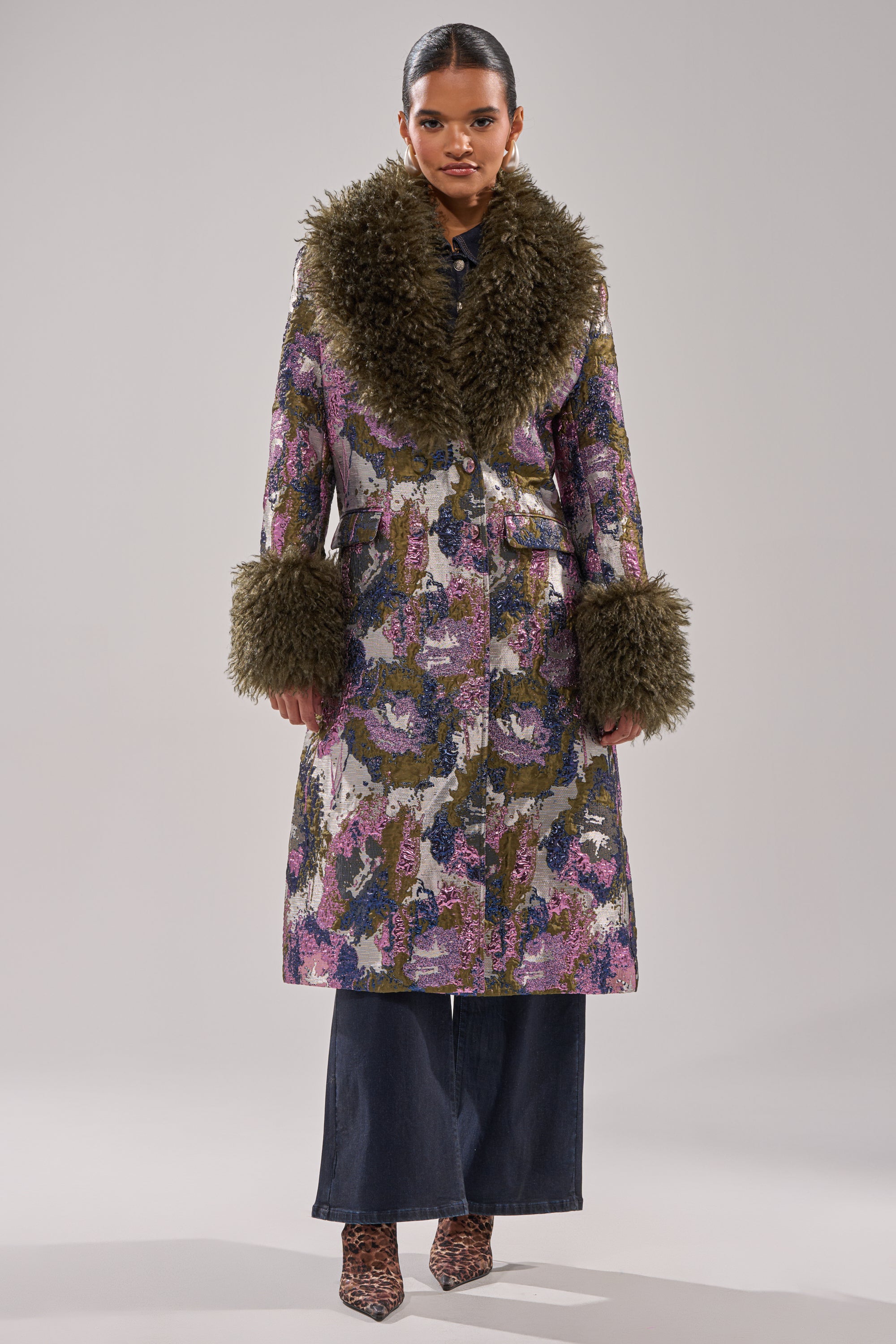 Wearing the GEORGINA BROCADE TRENCH with a large, fluffy faux fur trim in olive green, wide-leg pants, and patterned shoes, a person stands against a plain gray background.