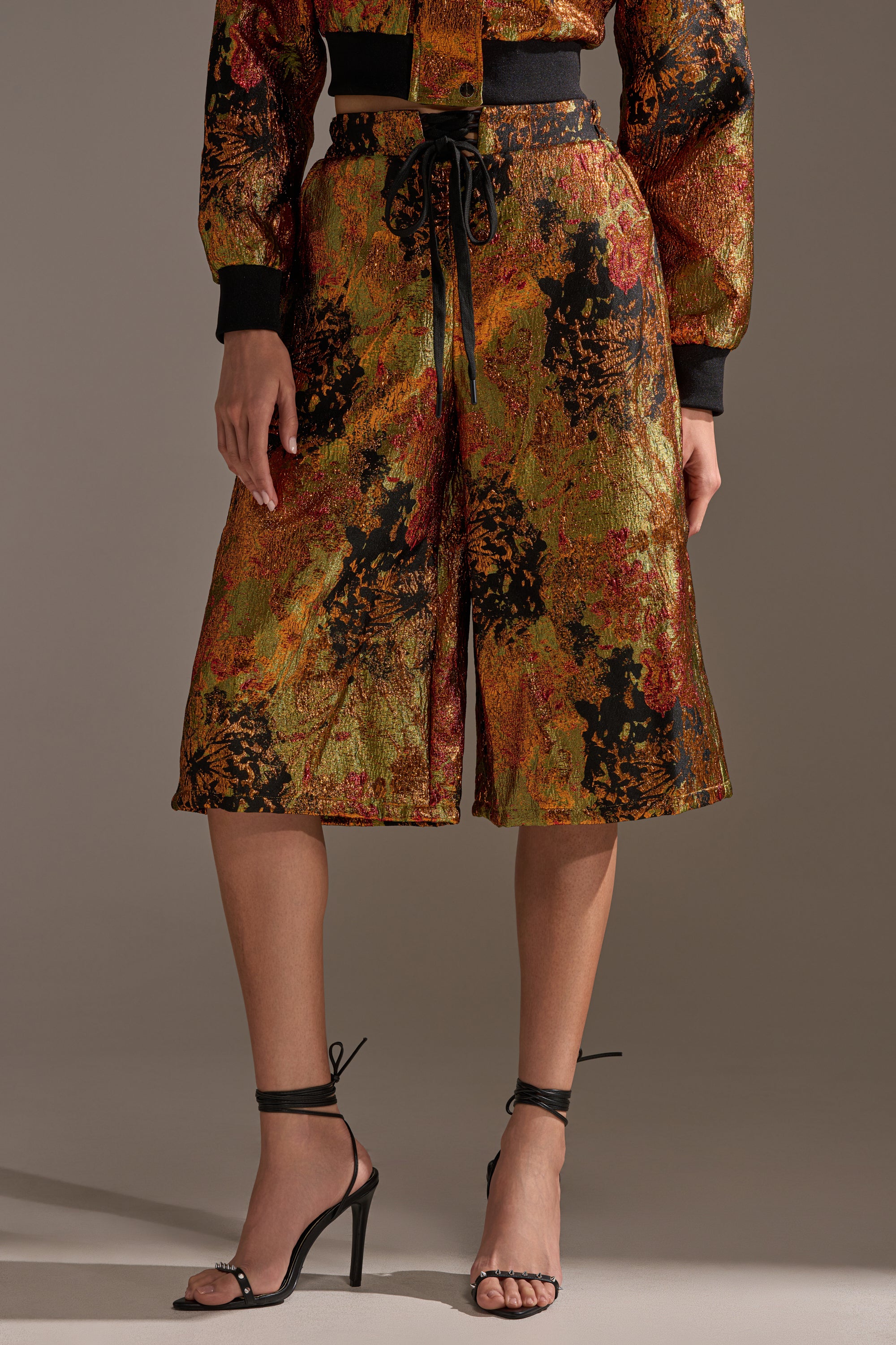 The lower body is shown wearing TWIN FLAME BROCADE BERMUDA SHORTS featuring gold, orange, black, and red patterns, paired with strappy black high-heeled sandals.