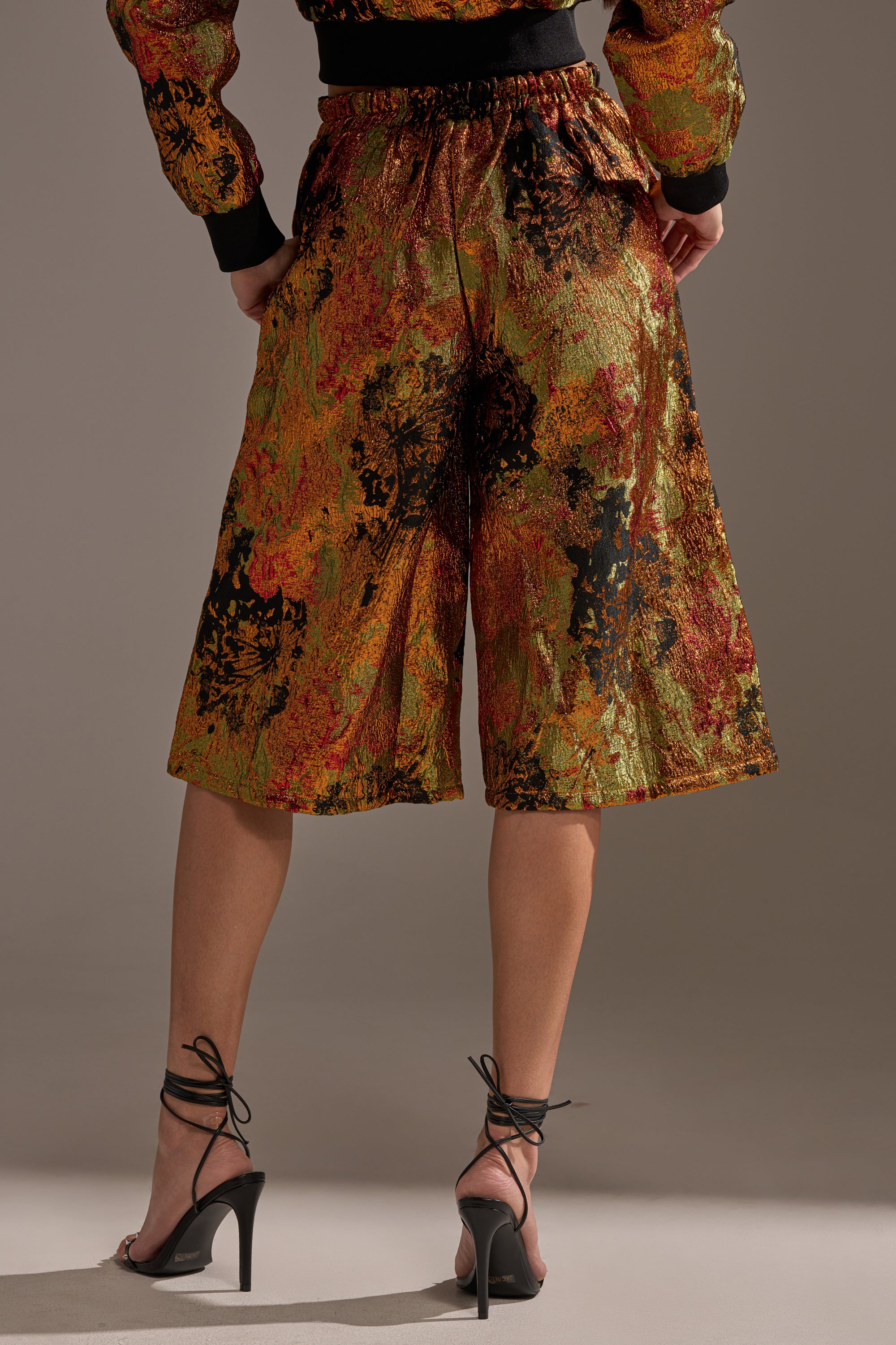 Someone wearing the TWIN FLAME BROCADE BERMUDA SHORTS, featuring an abstract multicolored orange, yellow, and black pattern, paired with strappy black high-heeled sandals; photo captured from behind.