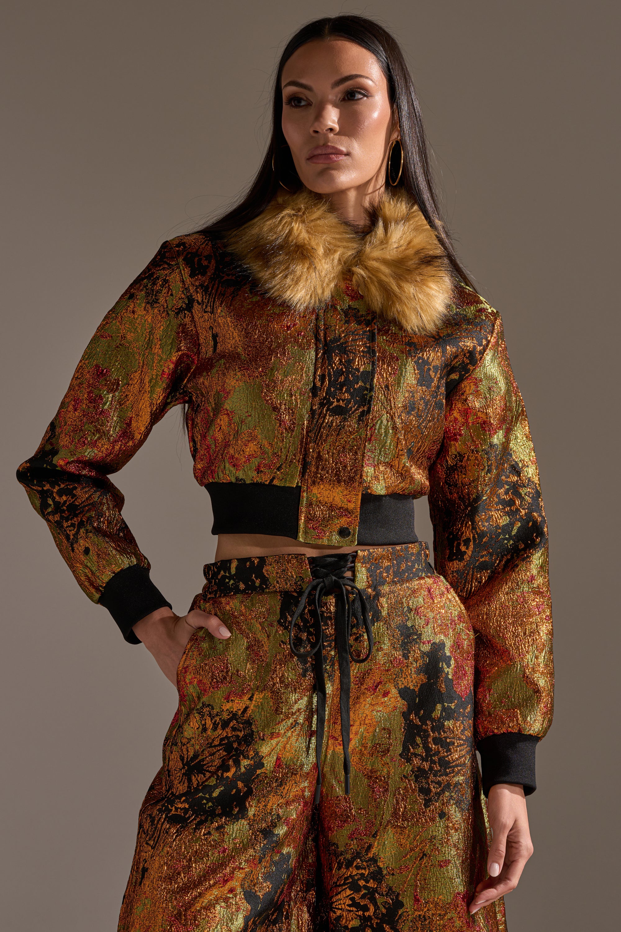 A woman stands confidently in the TWIN FLAME BROCADE BOMBER, a textured, multicolored jacket with a faux fur collar and cropped fit, paired with matching pants against a neutral background.